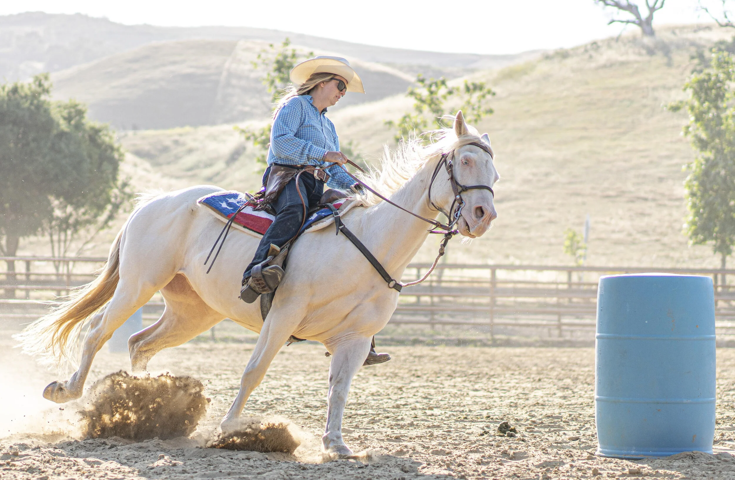 A person riding a white horse on a dirt arena, wearing a cowboy hat and sunglasses, going through a barrel racing course.