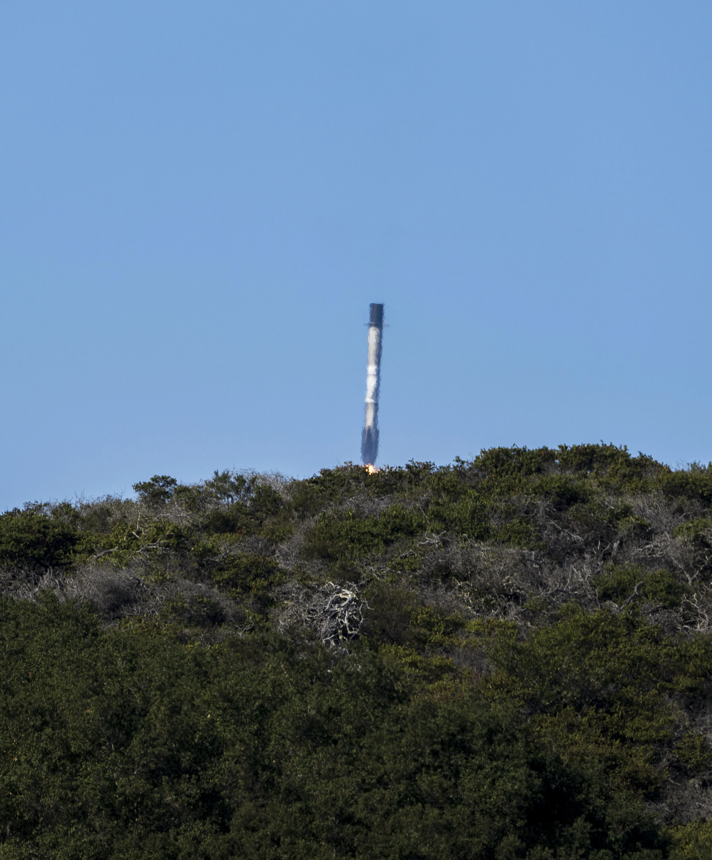 Rocket launching into the sky, with smoke at the bottom and a clear blue sky in the background, above a green treeline.