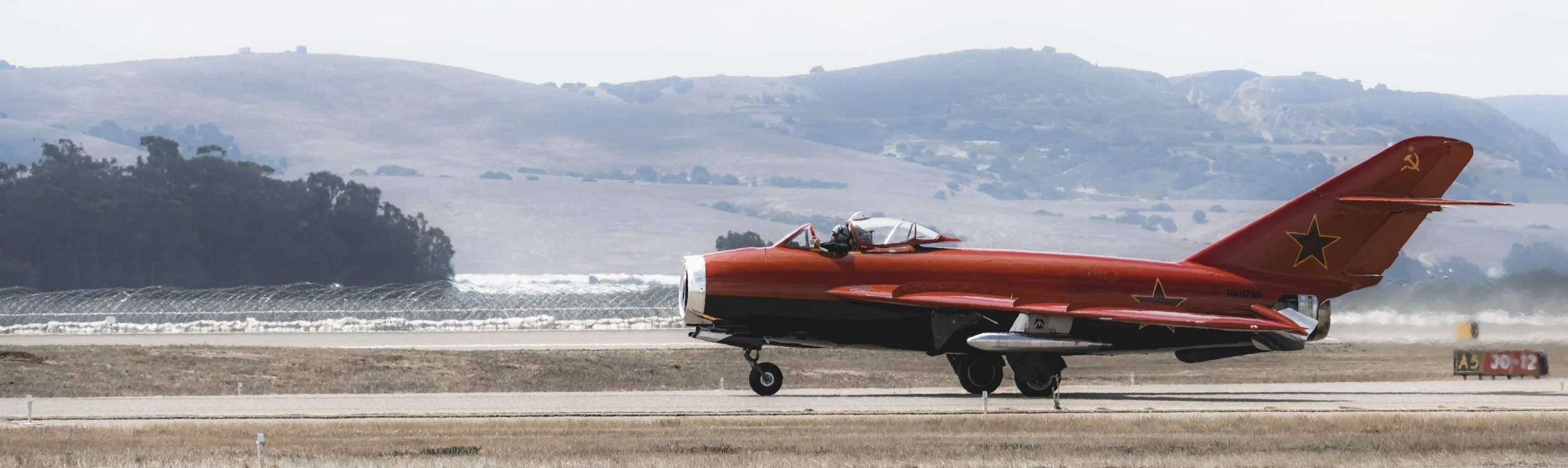A red fighter jet on the runway with a pilot inside. The jet has black stars and a hammer and sickle symbol on the tail.