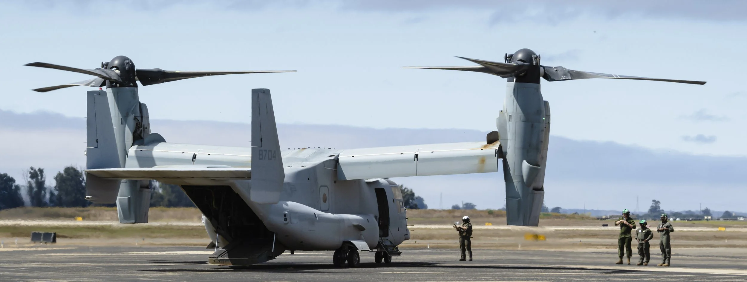 A military tiltrotor aircraft on a runway with soldiers nearby during the day.