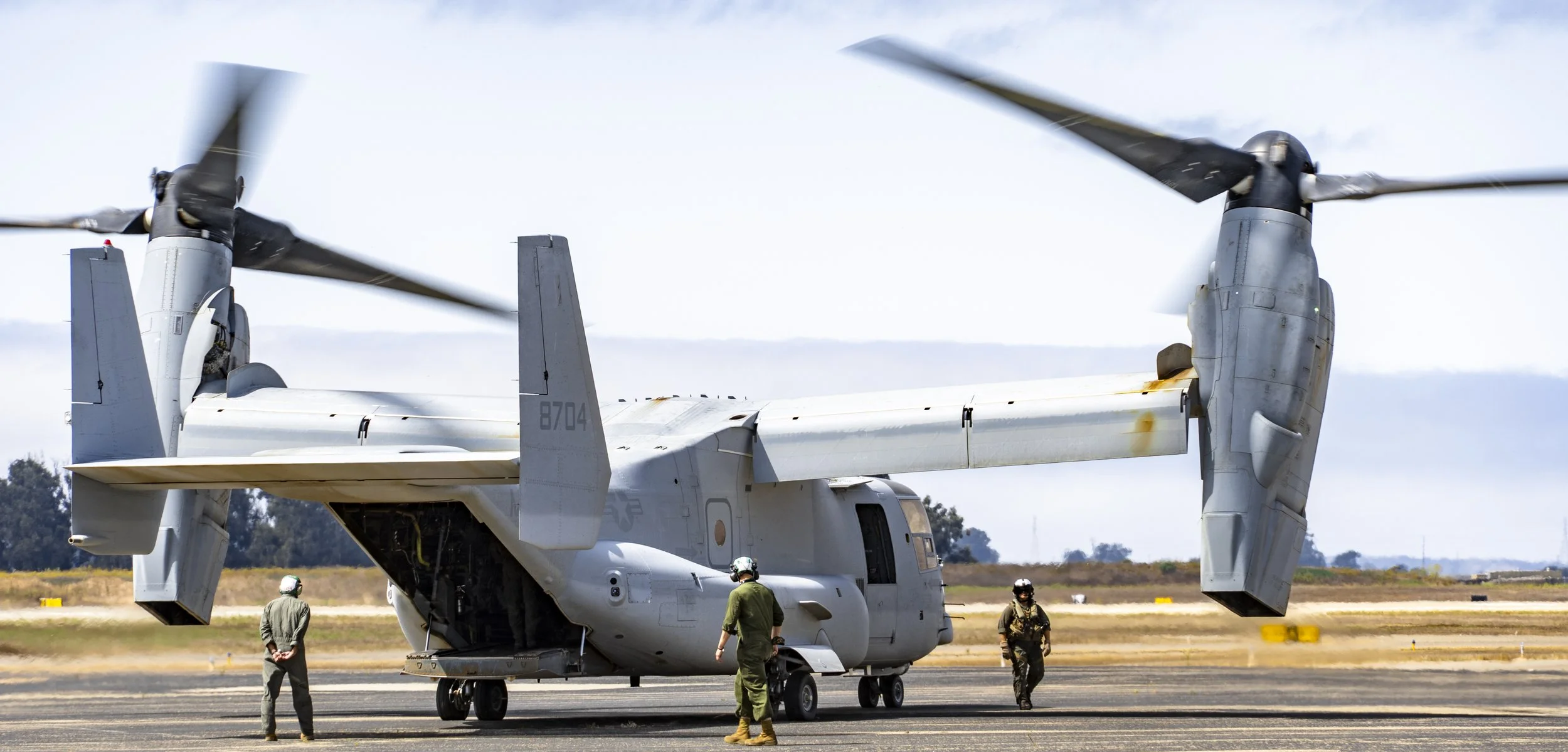 A military aircraft with a propeller rotor system on the ground, with personnel in military uniforms nearby at an airbase.