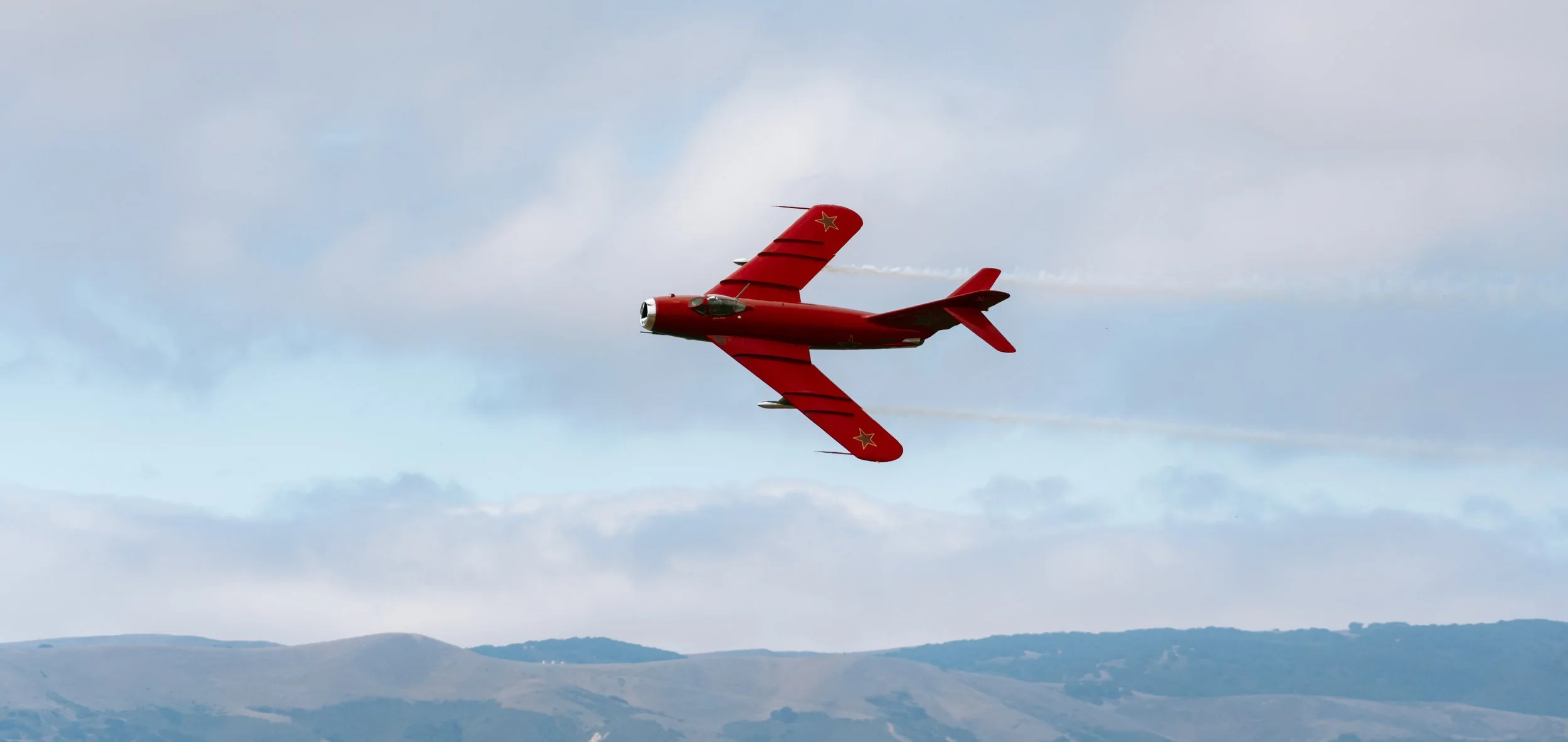 A red jet aircraft flying in the sky with hills in the background, leaving a trail of white smoke behind.