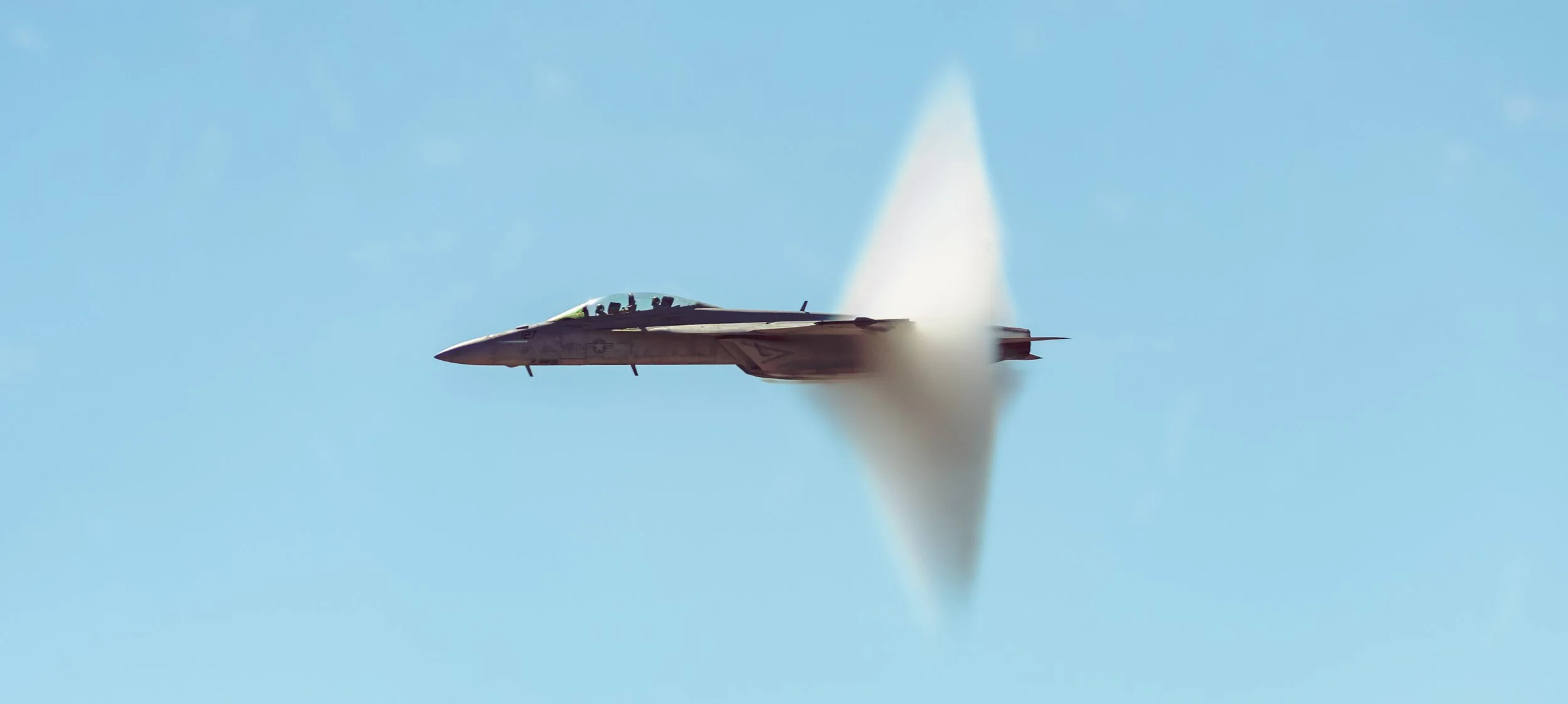 A fighter jet flying at high speed creating a vapor cone against a clear blue sky.