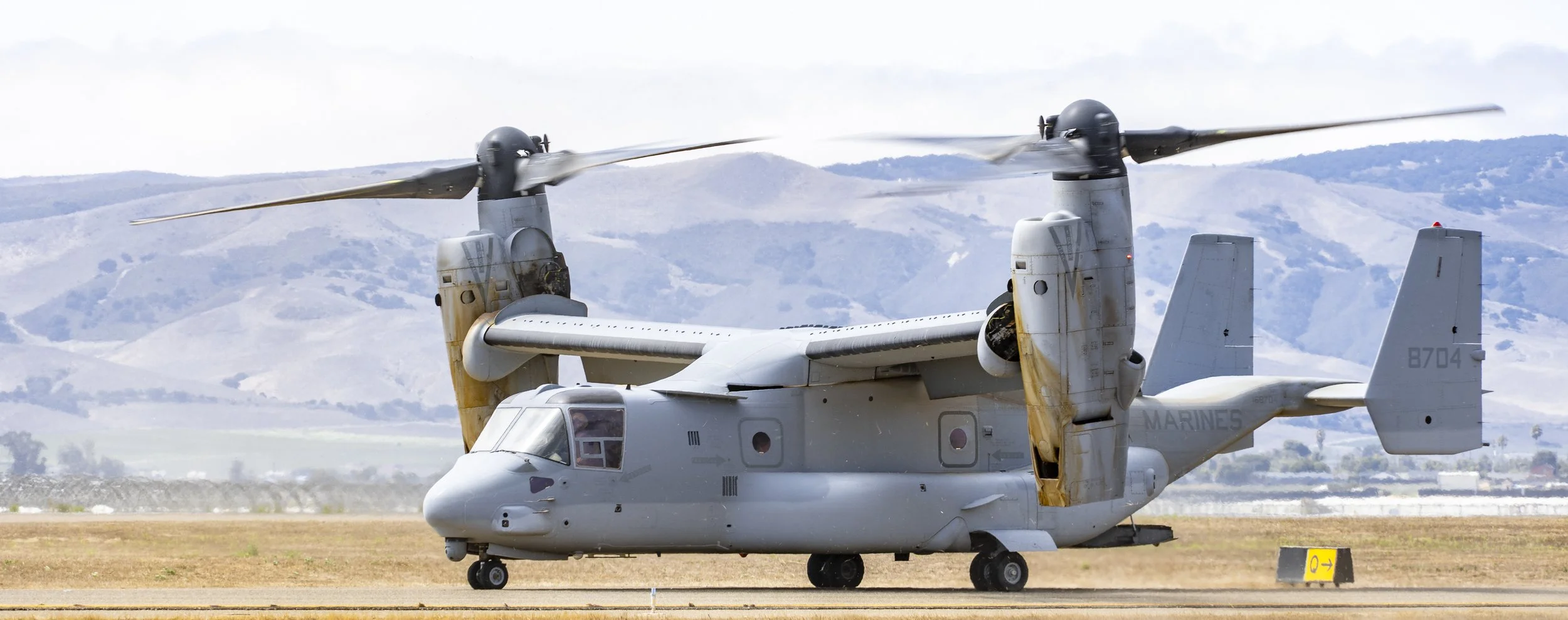 A Boeing V-22 Osprey military aircraft on the runway with its tiltrotors in vertical flight position, set against a background of rolling hills and cloudy sky.