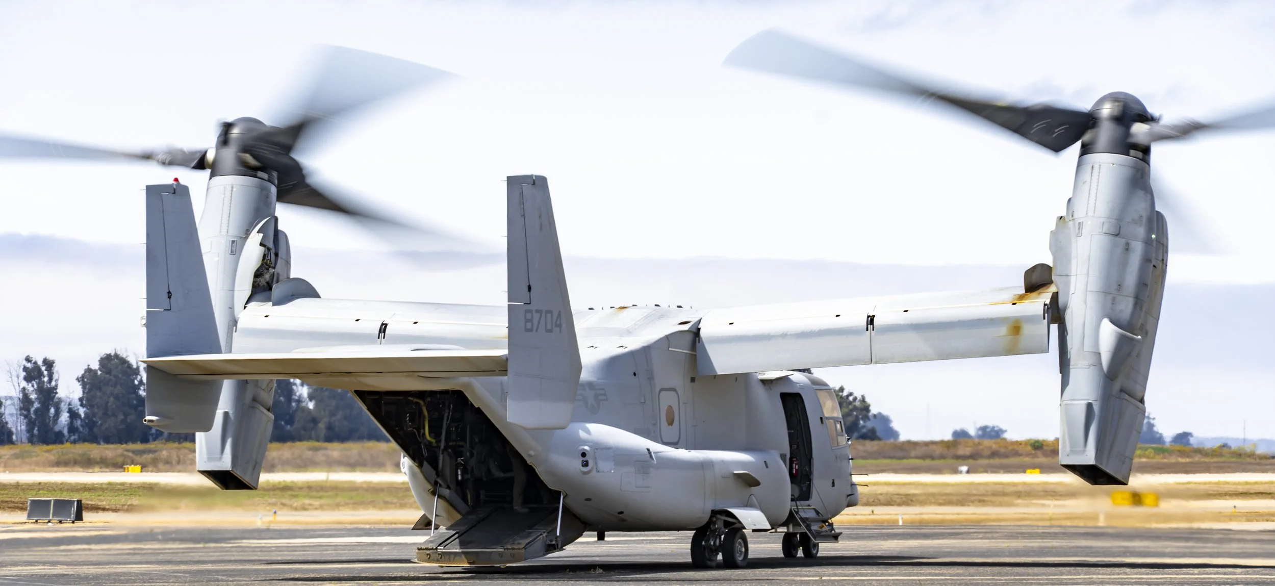 A military aircraft on the ground at an airfield, with its propellers spinning, and the sky and landscape in the background.