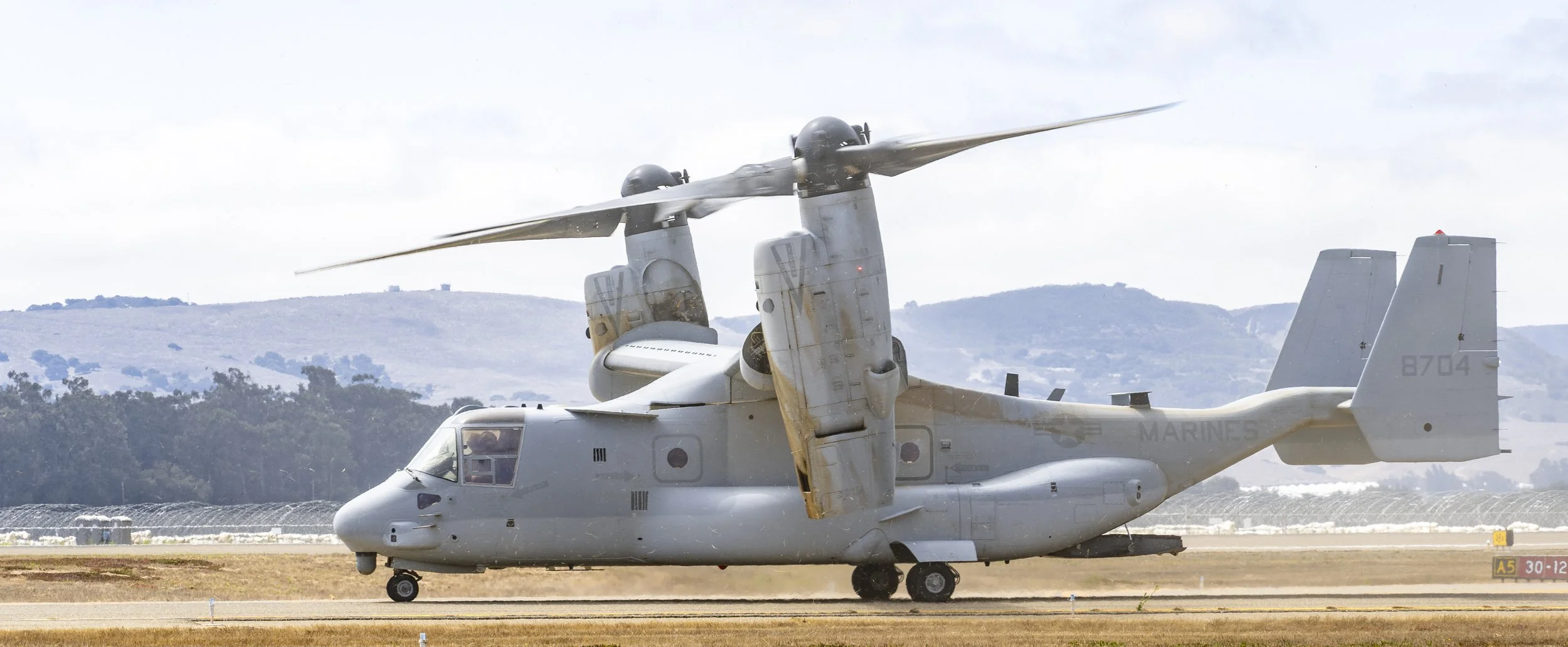 A military tiltrotor aircraft on the ground with its rotors in a vertical position, at an airfield with hills and trees in the background.