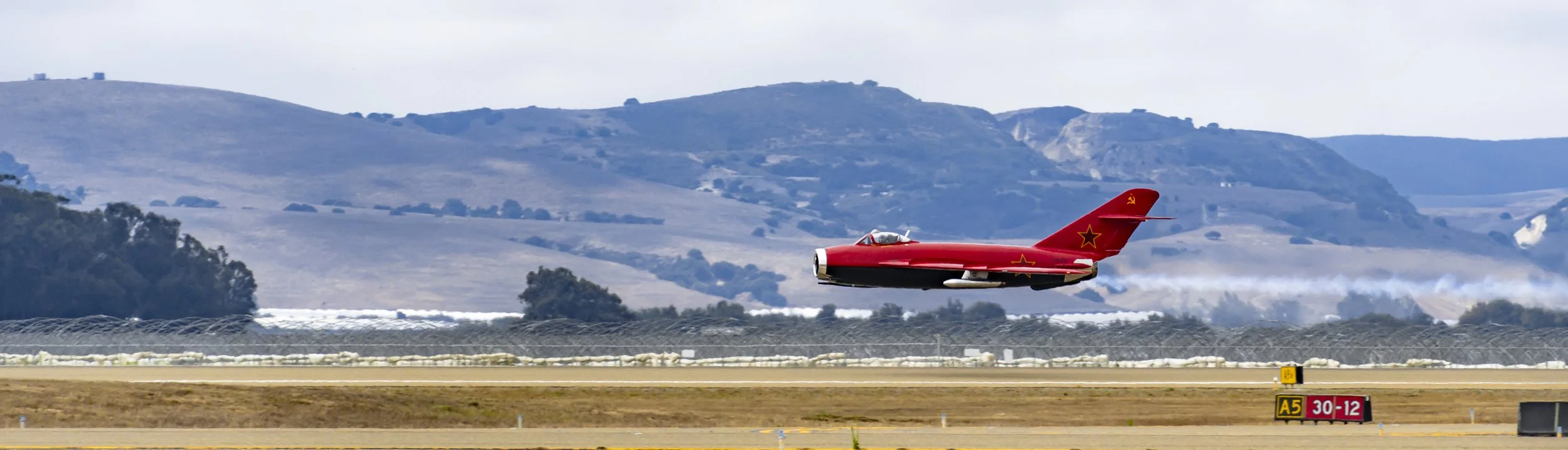 A red jet airplane with Soviet markings takes off from a runway with mountains in the background.