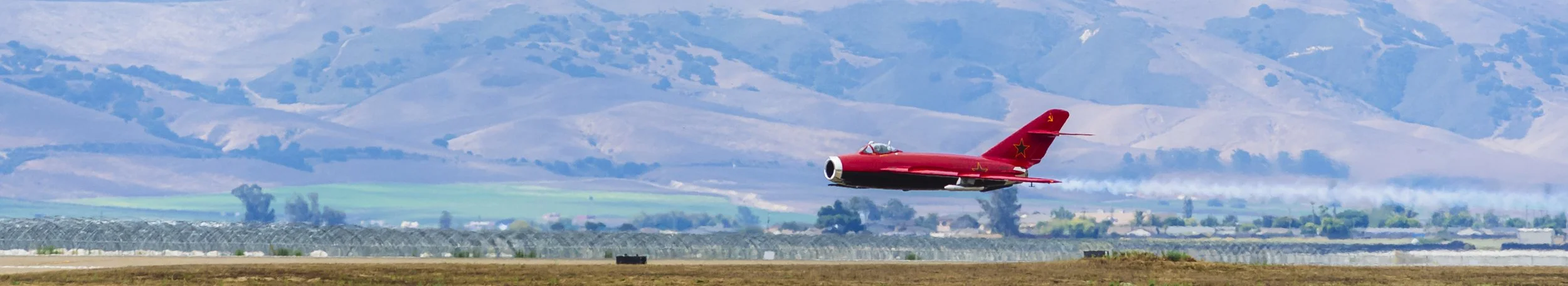 A red fighter jet soaring over a flat landscape with rolling hills in the background.