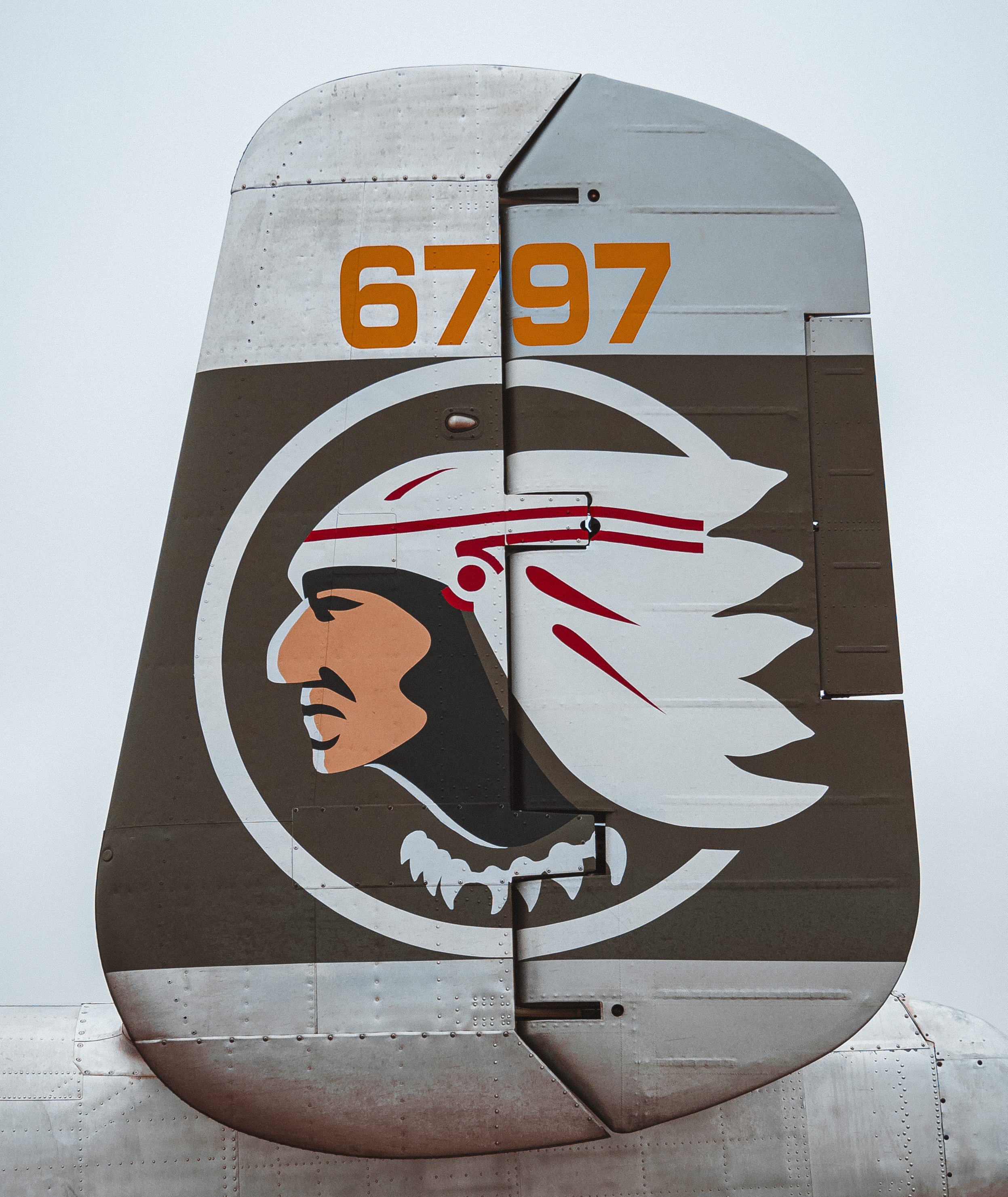 Close-up of an aircraft tail fin displaying a stylized Native American profile with a feathered headdress and the number 6797 in orange on a gray background.