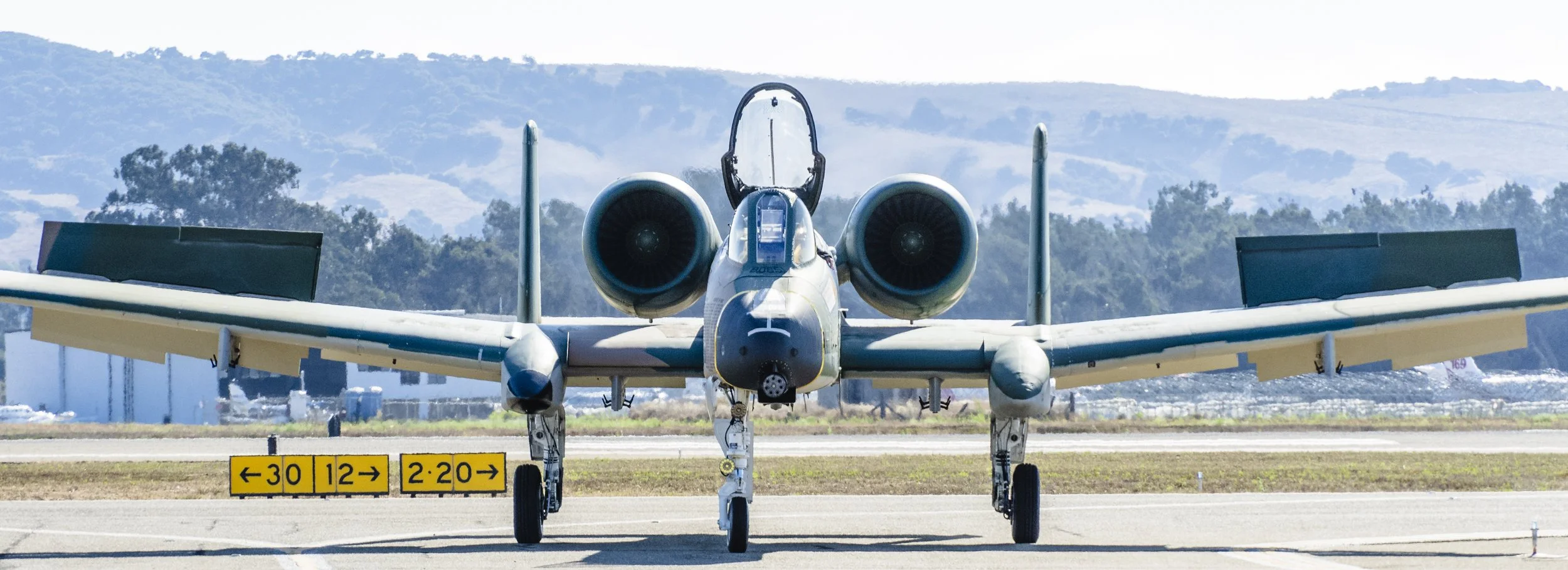 Front view of a fighter jet on the runway, with trees and hills in the background and directional signs on the ground.