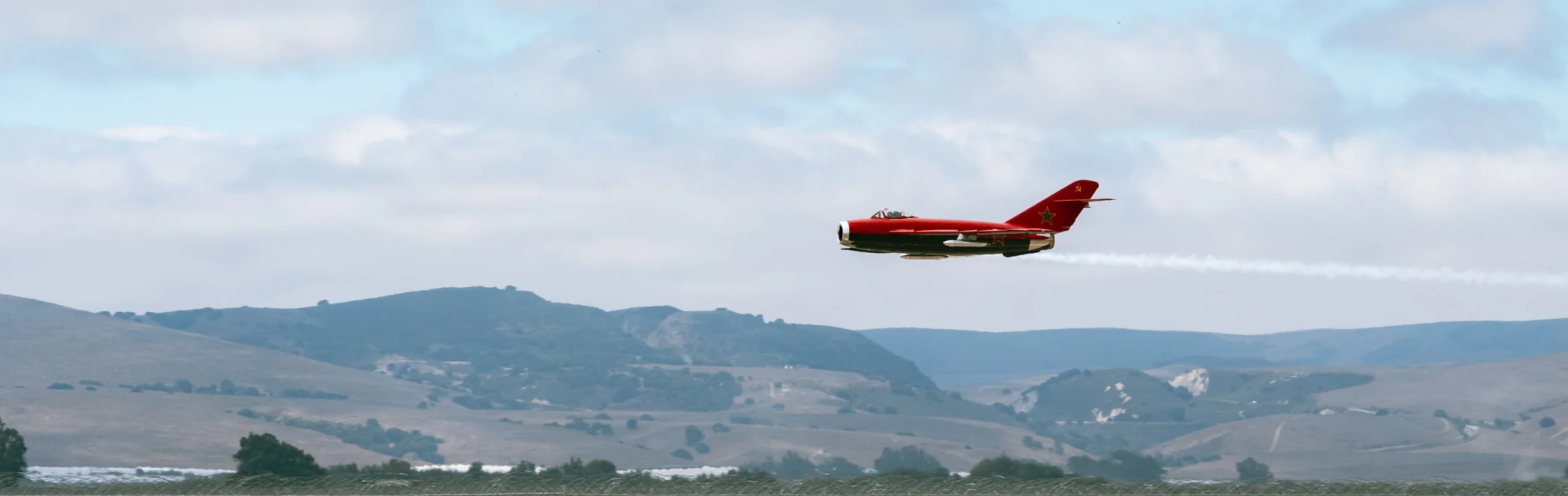 Red jet aircraft flying over a landscape with hills and scattered trees, leaving a white smoke trail behind.