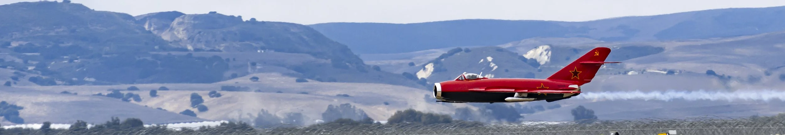 A red vintage jet with a Soviet star emblem is flying over a landscape of rolling hills and farmland.