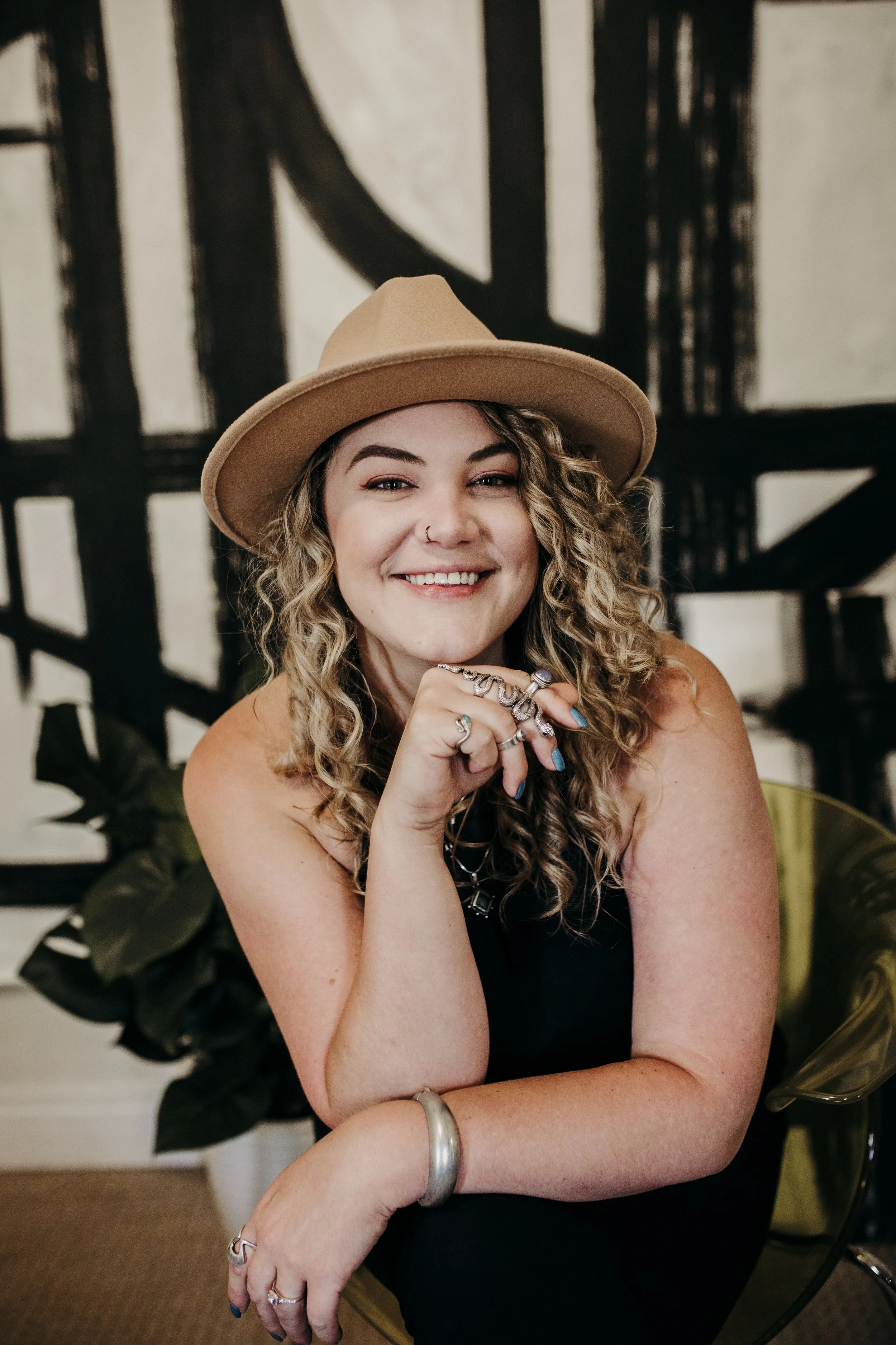 A smiling woman with curly blonde hair wearing a tan wide-brimmed hat, black top, and multiple rings on her fingers, sitting indoors with a large plant and a geometric black and white wall in the background.