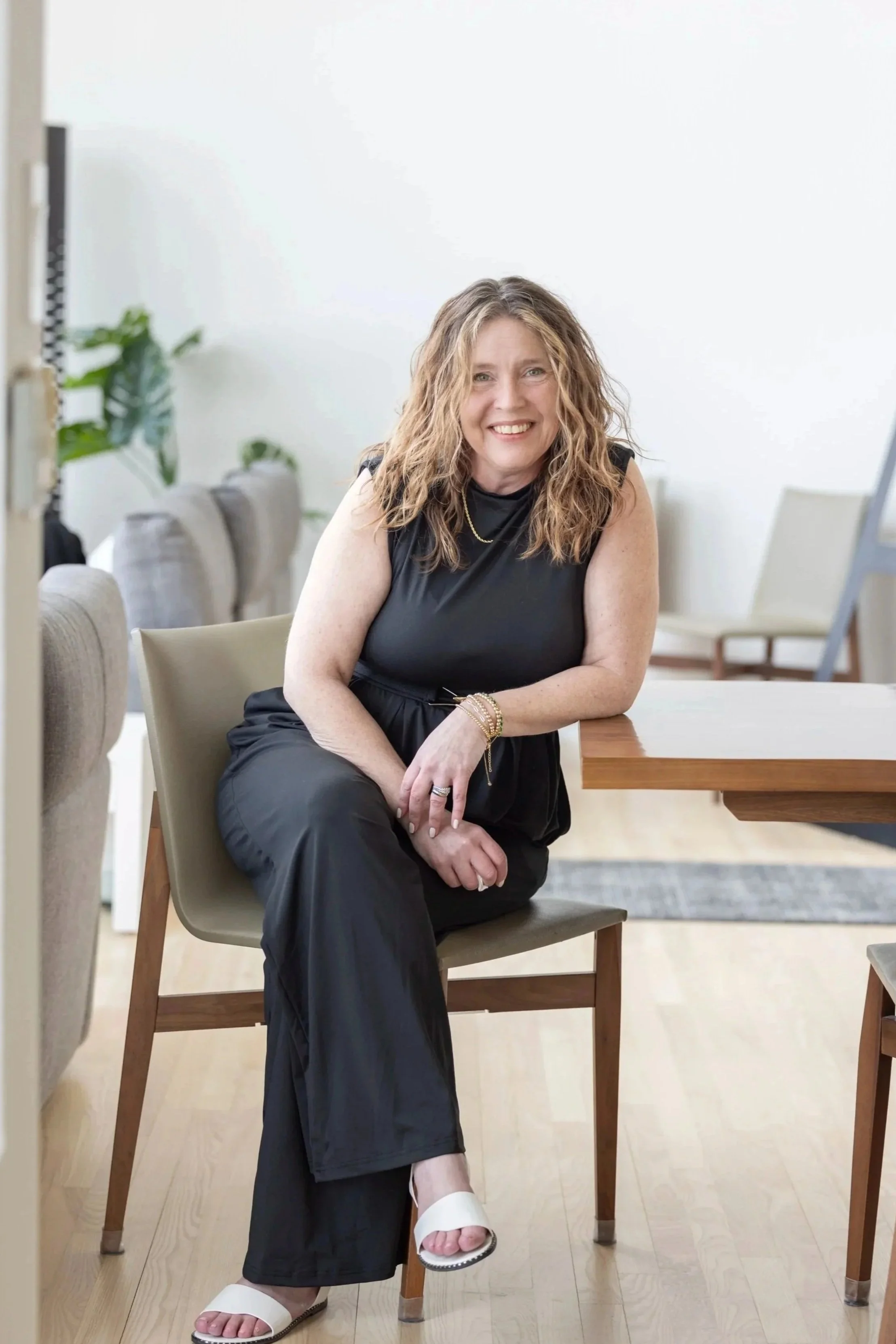 A woman with wavy, shoulder-length hair wearing a black sleeveless top and black pants, sitting on a chair at a wooden table in a bright room with minimalist decor and potted plants.