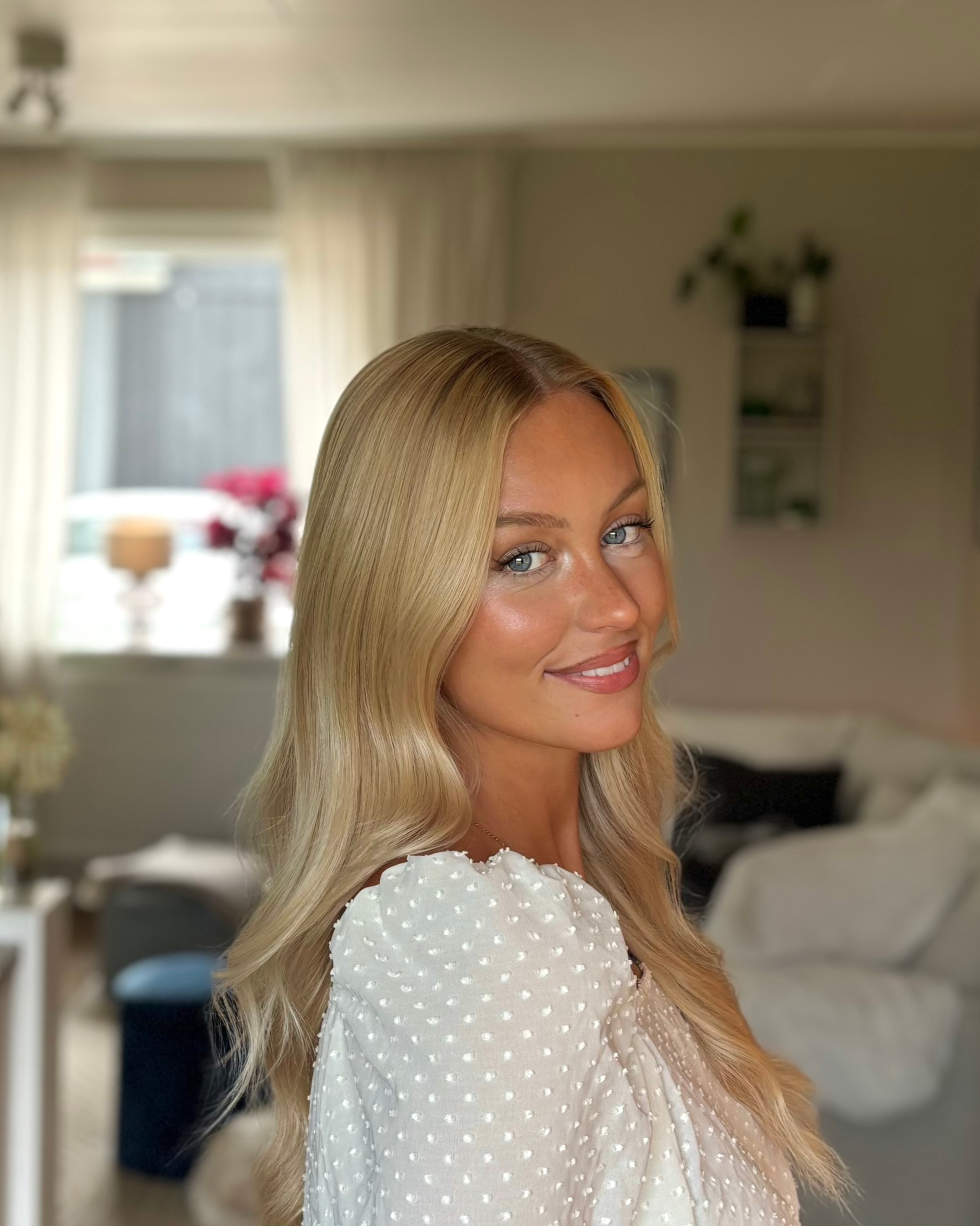 A woman with blonde wavy hair and light blue eyes smiling in a well-lit living room with beige curtains and furniture.