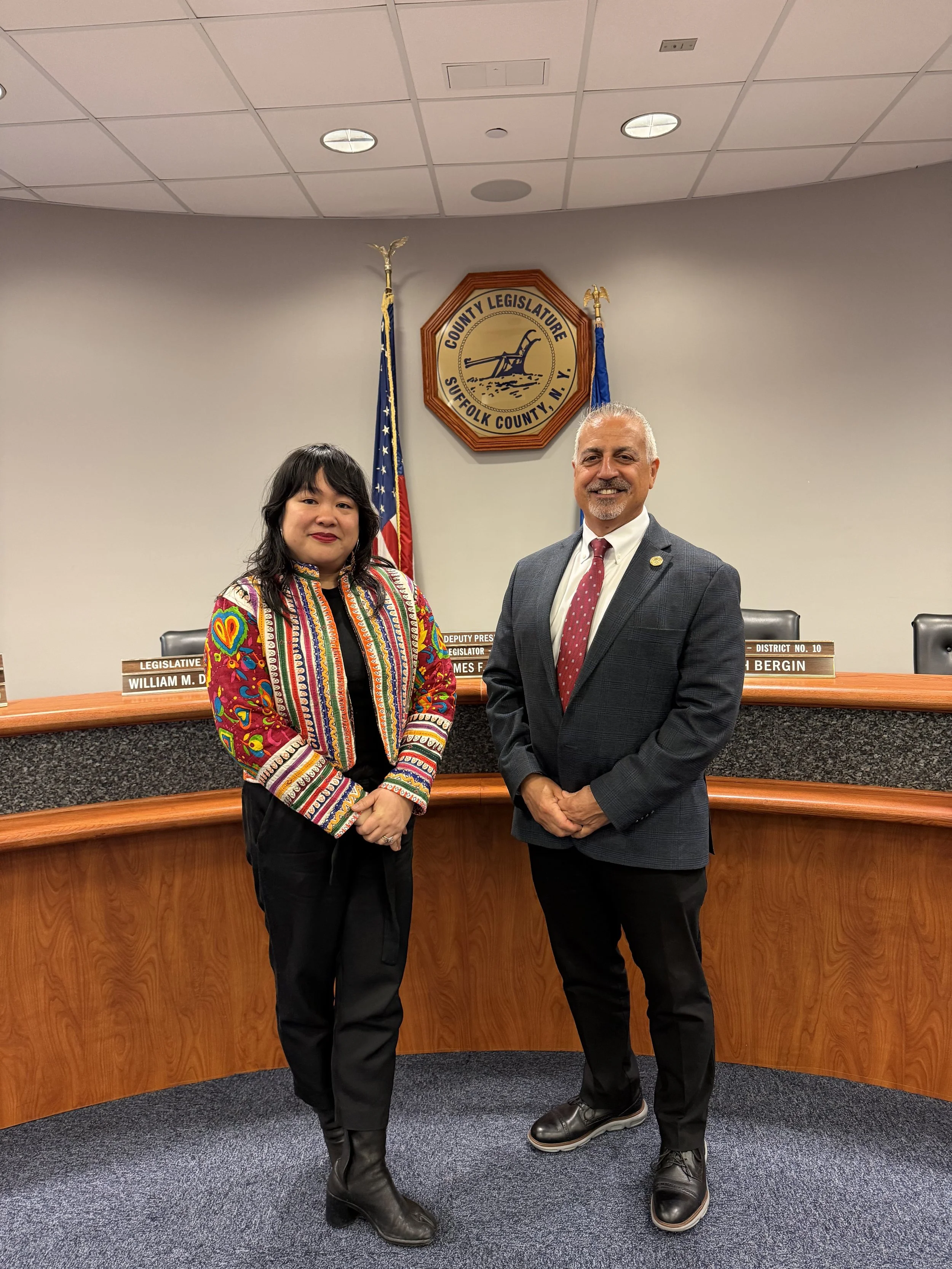 Artist Jessamyn Go is standing on the left whearing a color ful jacket and black pants hands crossed in front of her.  Standing to here right James Mazzarella is wearing a red tie and blazer.  Standing in front of a wooden bureau and 2 American flag