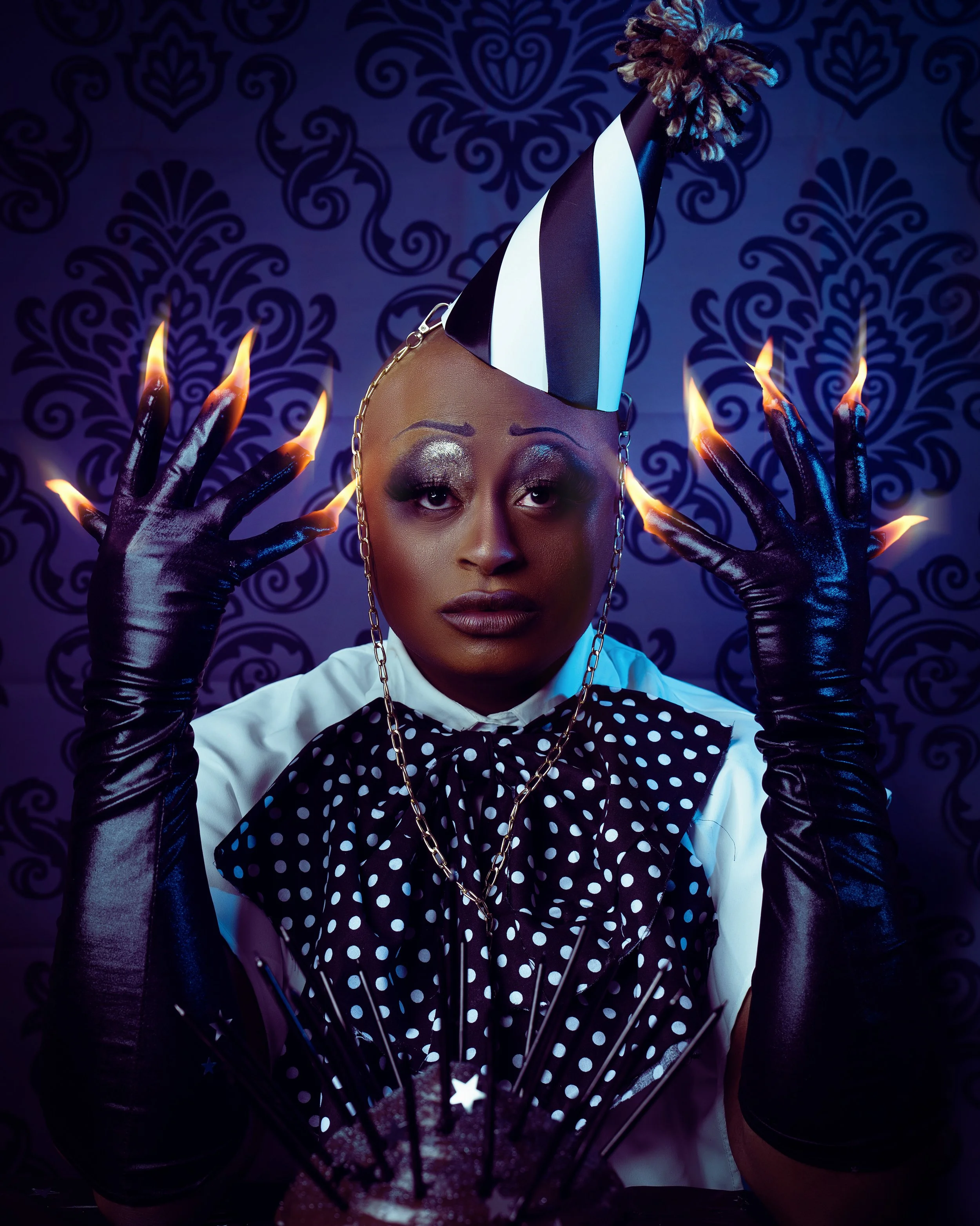 Self-portrait in a striped dunce cap with a gold chain, black leather styling, fire at the fingertips, high mime makeup, baroque backdrop with a giant cake — KEN X KEEN Orlando