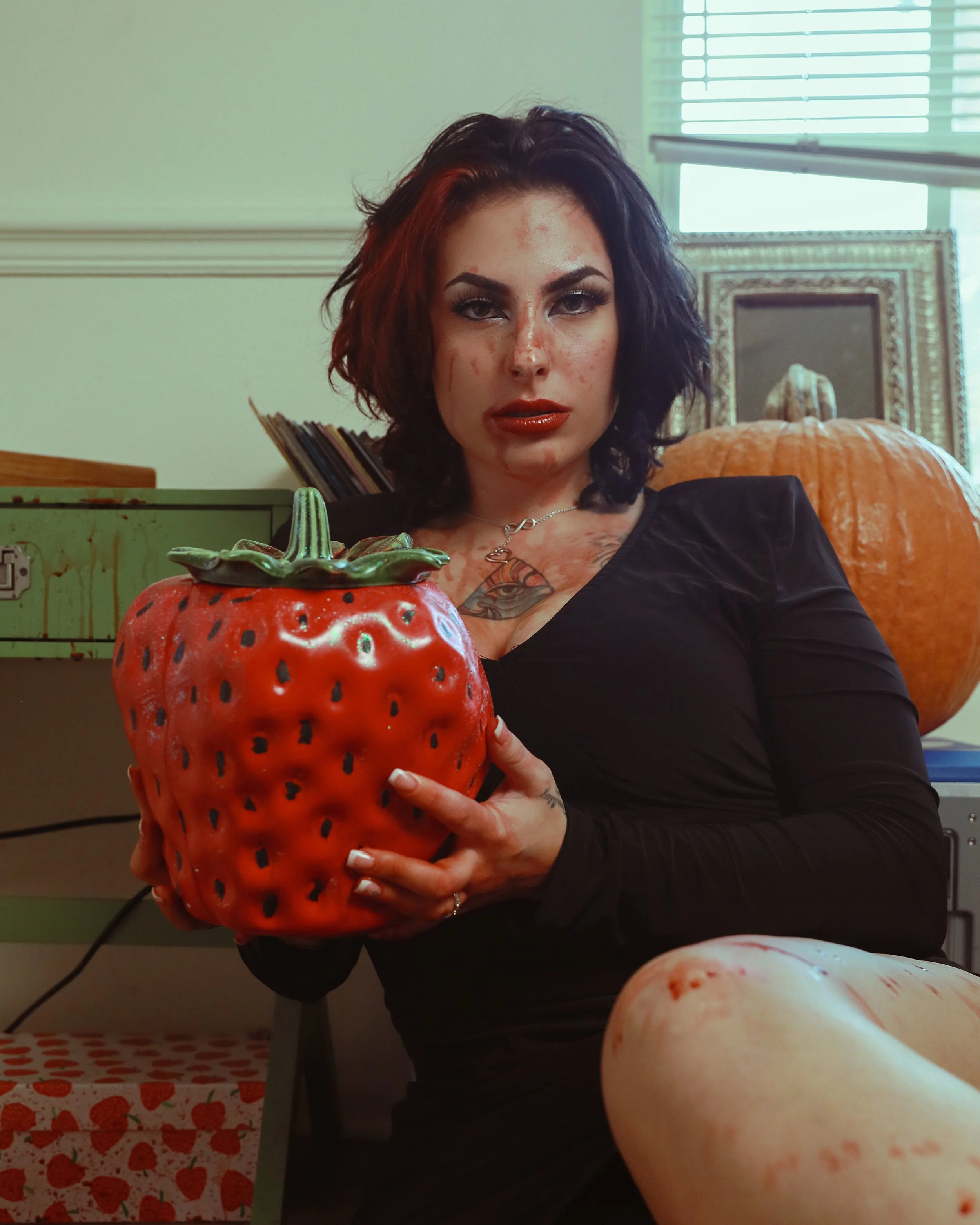 Model with dark smudged makeup and faux blood detailing, holding an oversized decorative strawberry beside a carved pumpkin — horror-glam editorial, KEN X KEEN Orlando