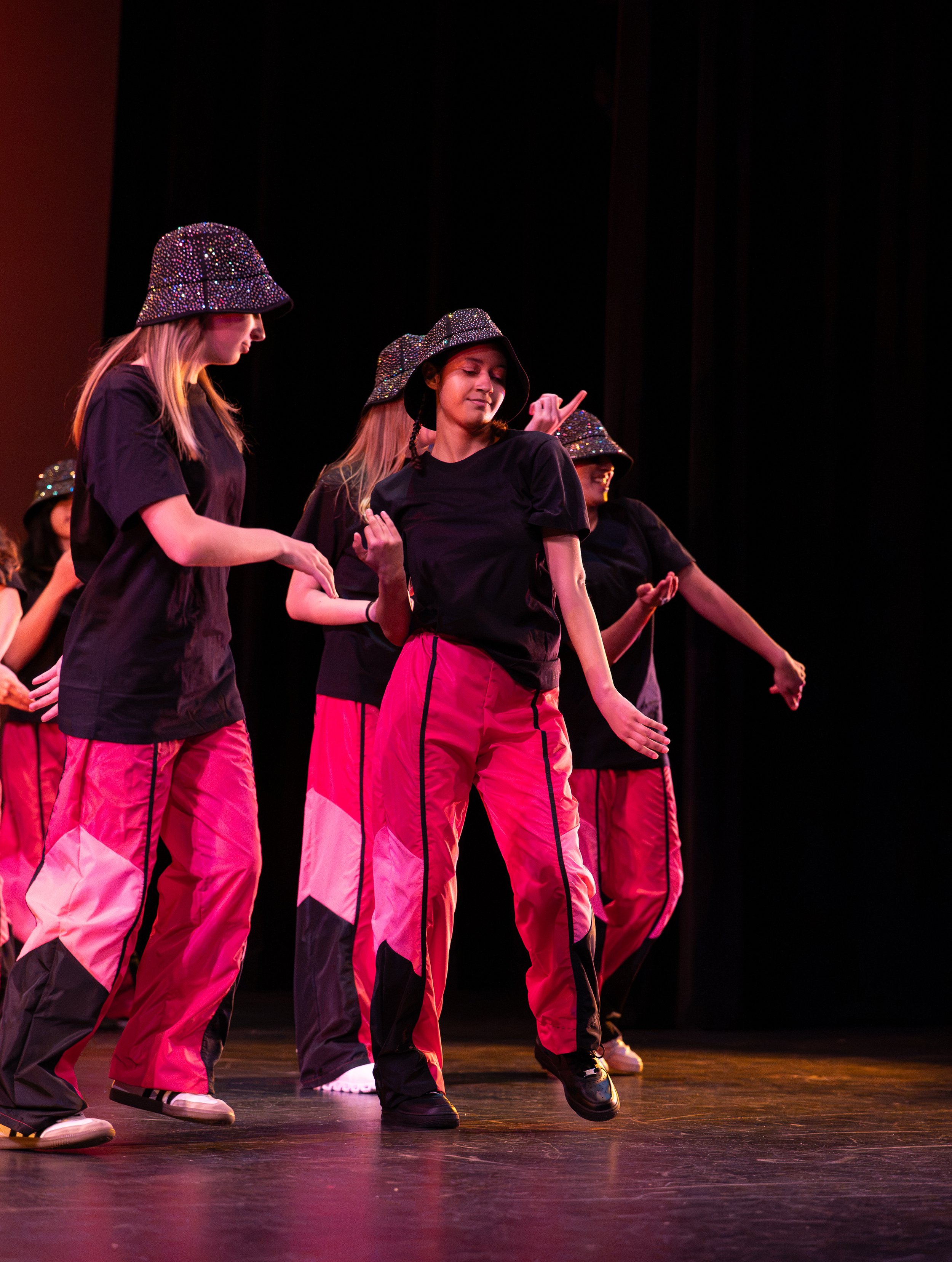 Group of young pop styles danciners on stage, wearing matching black t-shirts, pink and black track pants, and glittery bucket hats, with a dark background and stage lighting.