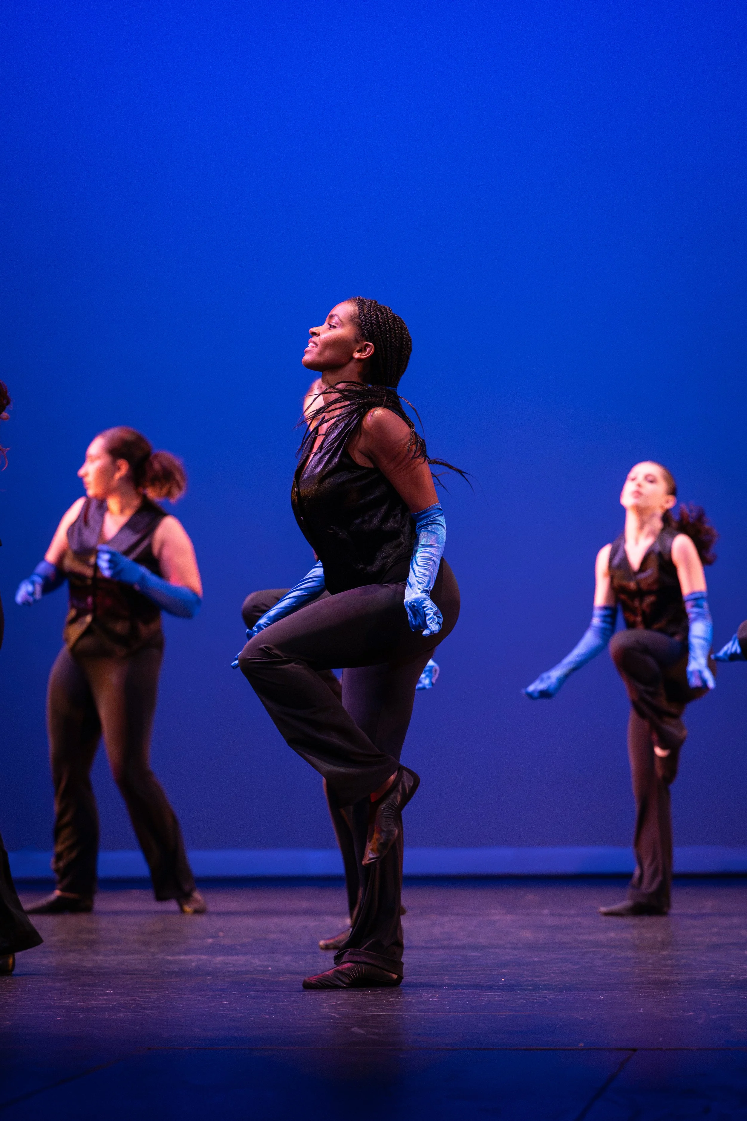 Group of women in dance costumes wearing black outfits and blue gloves performing a jazz dance on stage with a blue background.