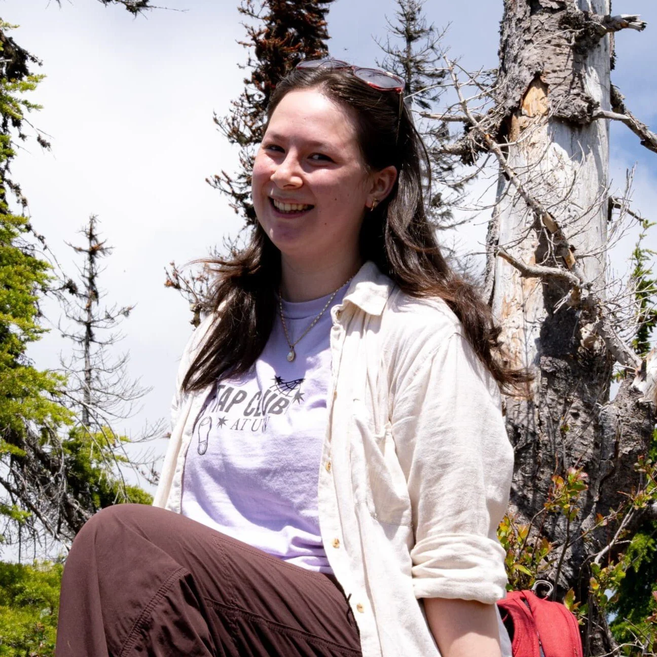 A young woman sitting outdoors in front of a weathered tree with green and brown foliage, smiling at the camera on a partly cloudy day.
