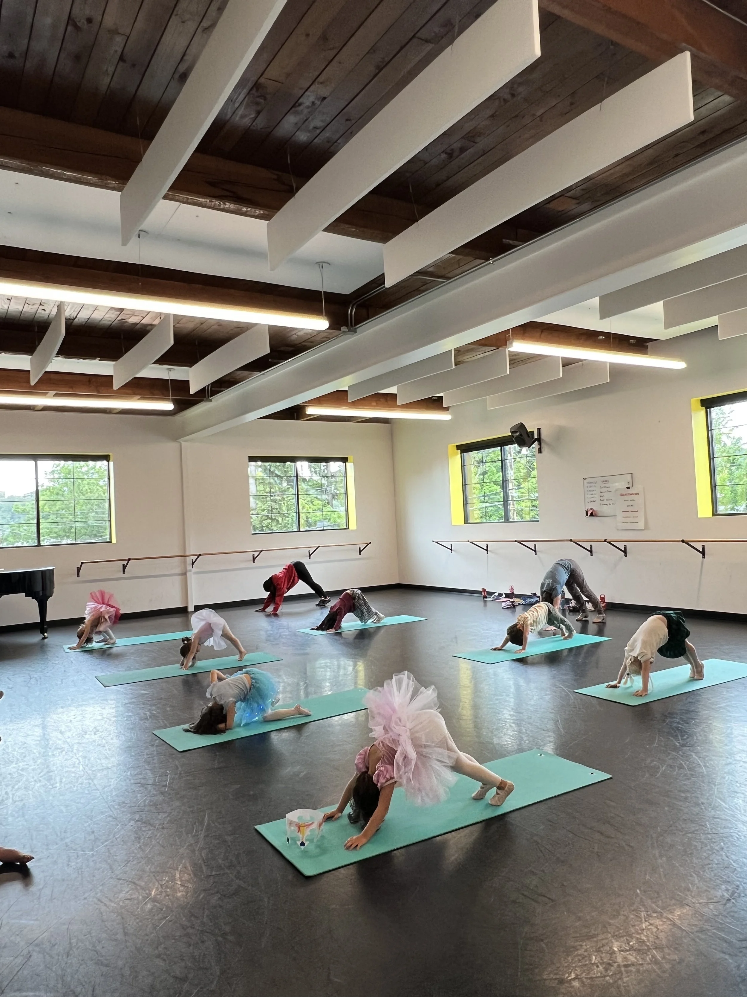 Children practicing yoga poses on mats in a dance studio with wooden floors and large windows.