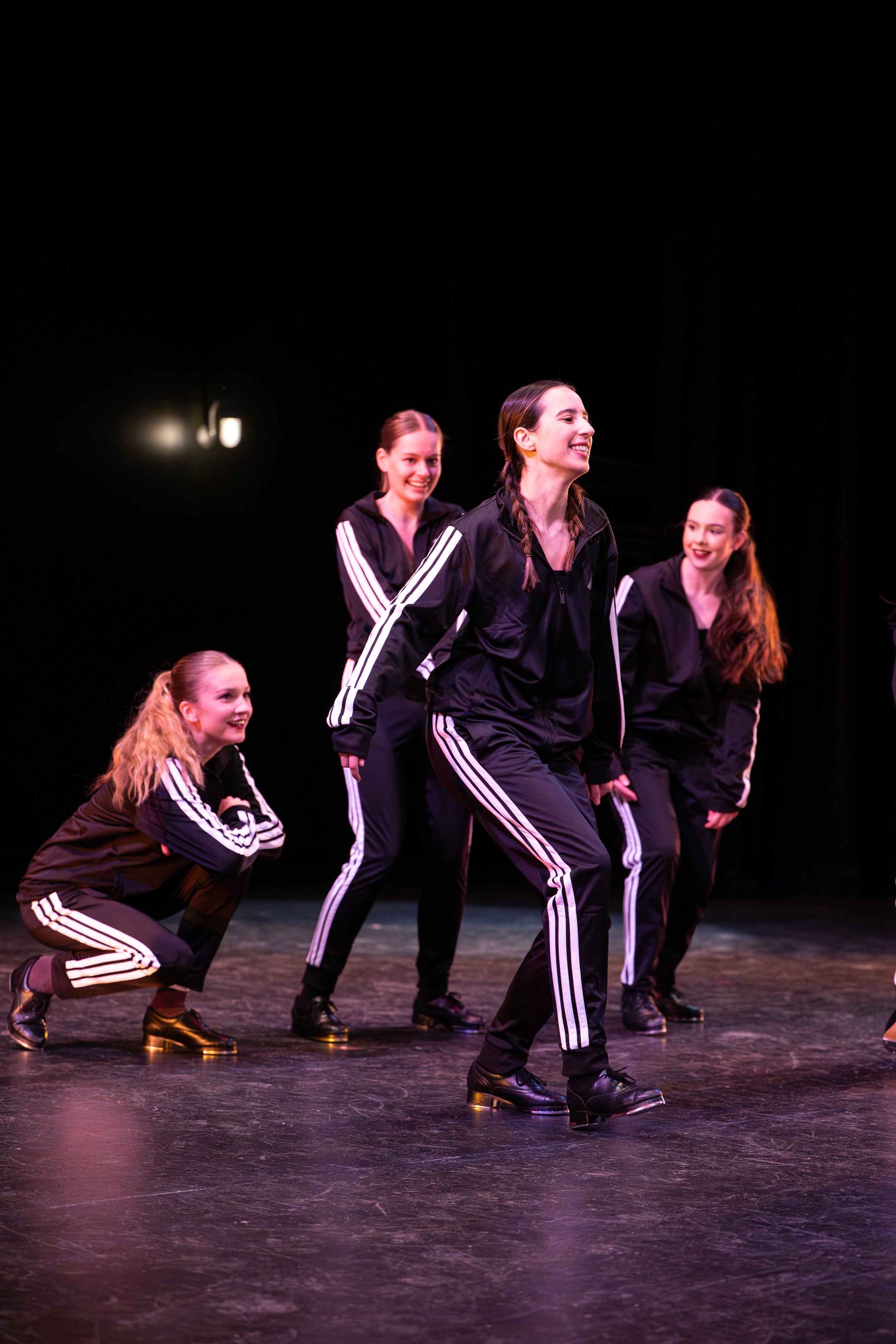 Group of young women performing a tap dance routine on stage, wearing black tracksuits with white stripes and black tap shoes