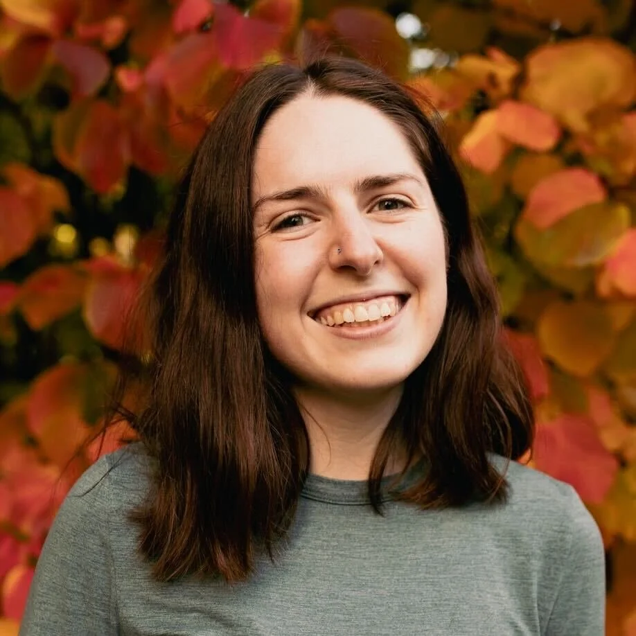 A young woman with shoulder-length brown hair smiling, wearing a black top, standing outdoors with a fall leaf background.