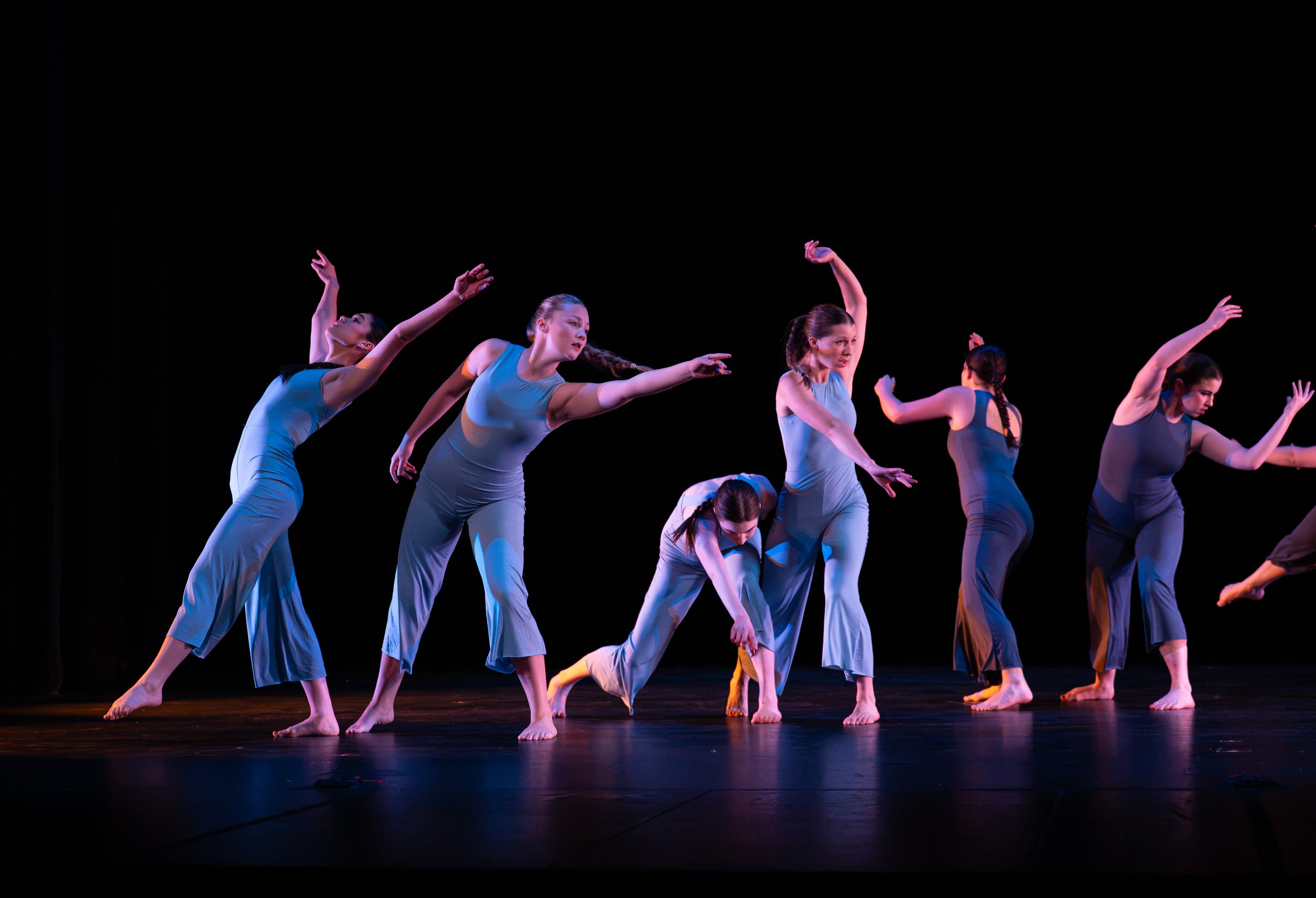 A group of modern dancers performing on stage, dressed in matching gray costumes, with some bending forward and others extending their arms, against a black background.