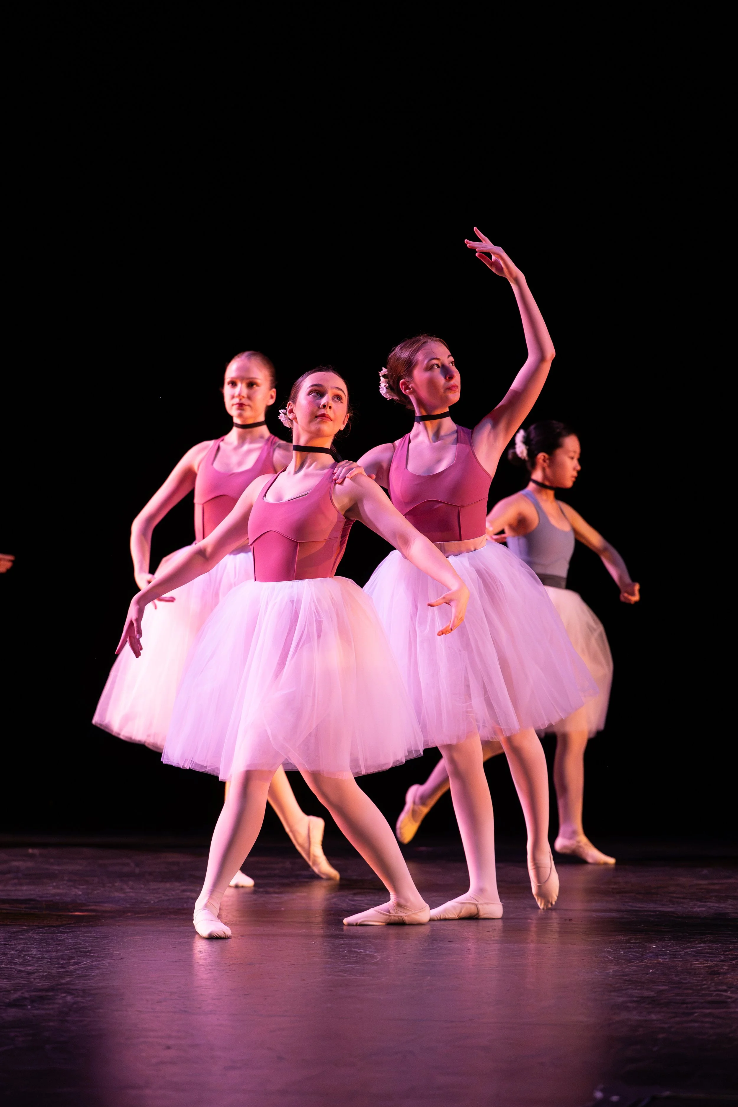 Four ballet dancers dressed in pink tutus and black choker necklaces performing on a stage with a black background, captured mid-dance.