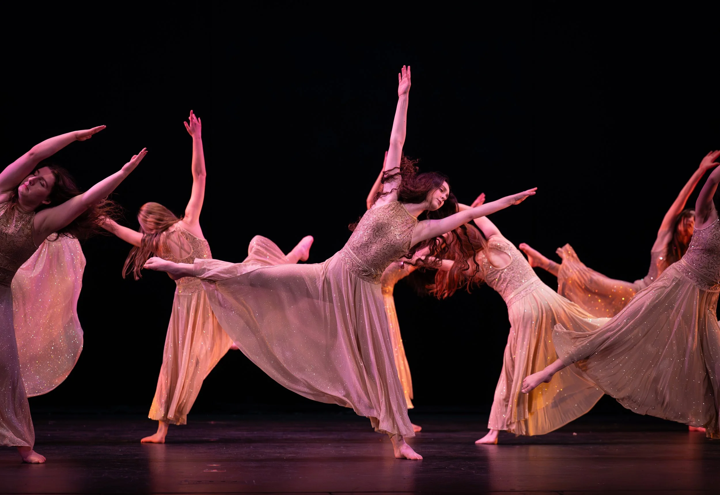 A group of lyrical dancers performing to music on stage, wearing golden costumes and in a tilted balancing shape.