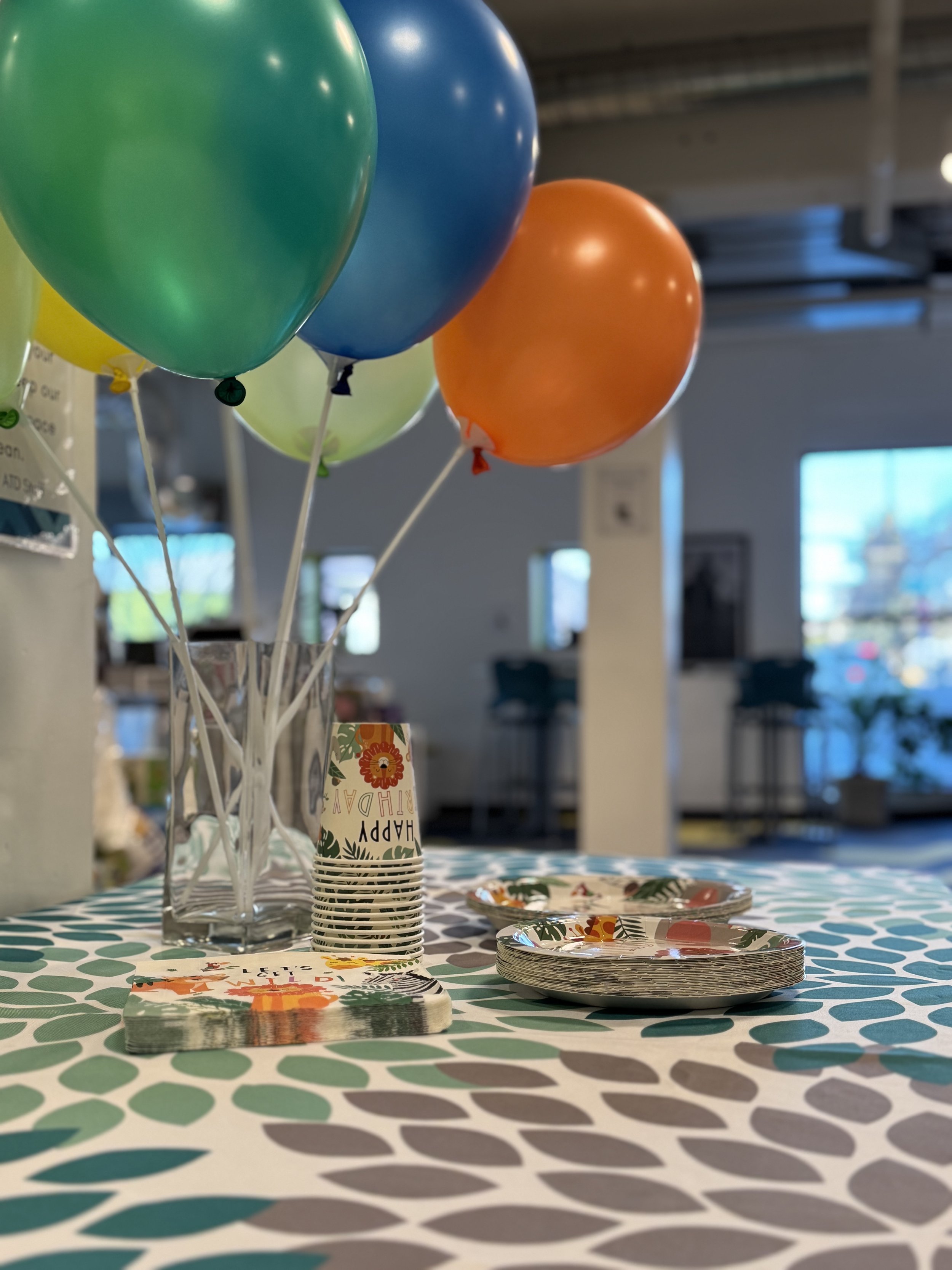 Colorful balloons tied to a clear vase on a decorated table with plates and cups.