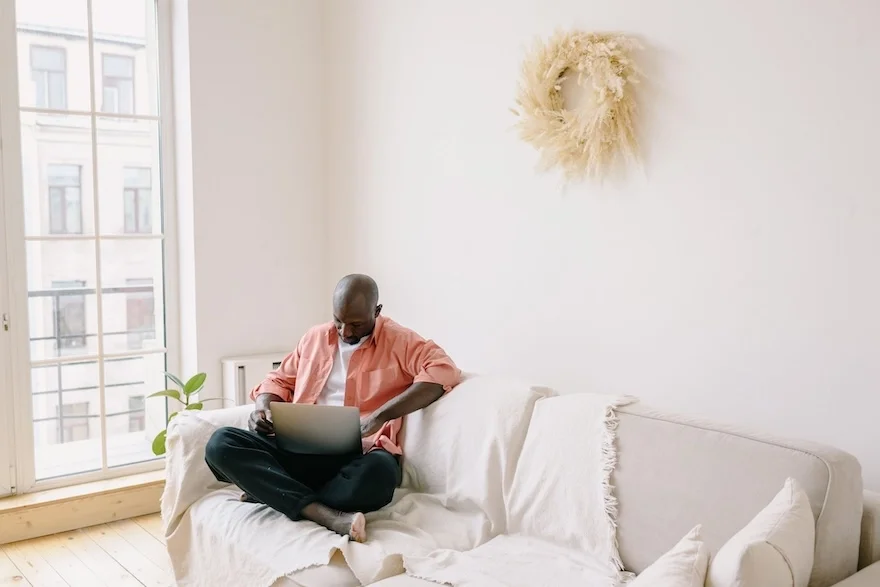 Man on a couch by a window in a white room at a computer