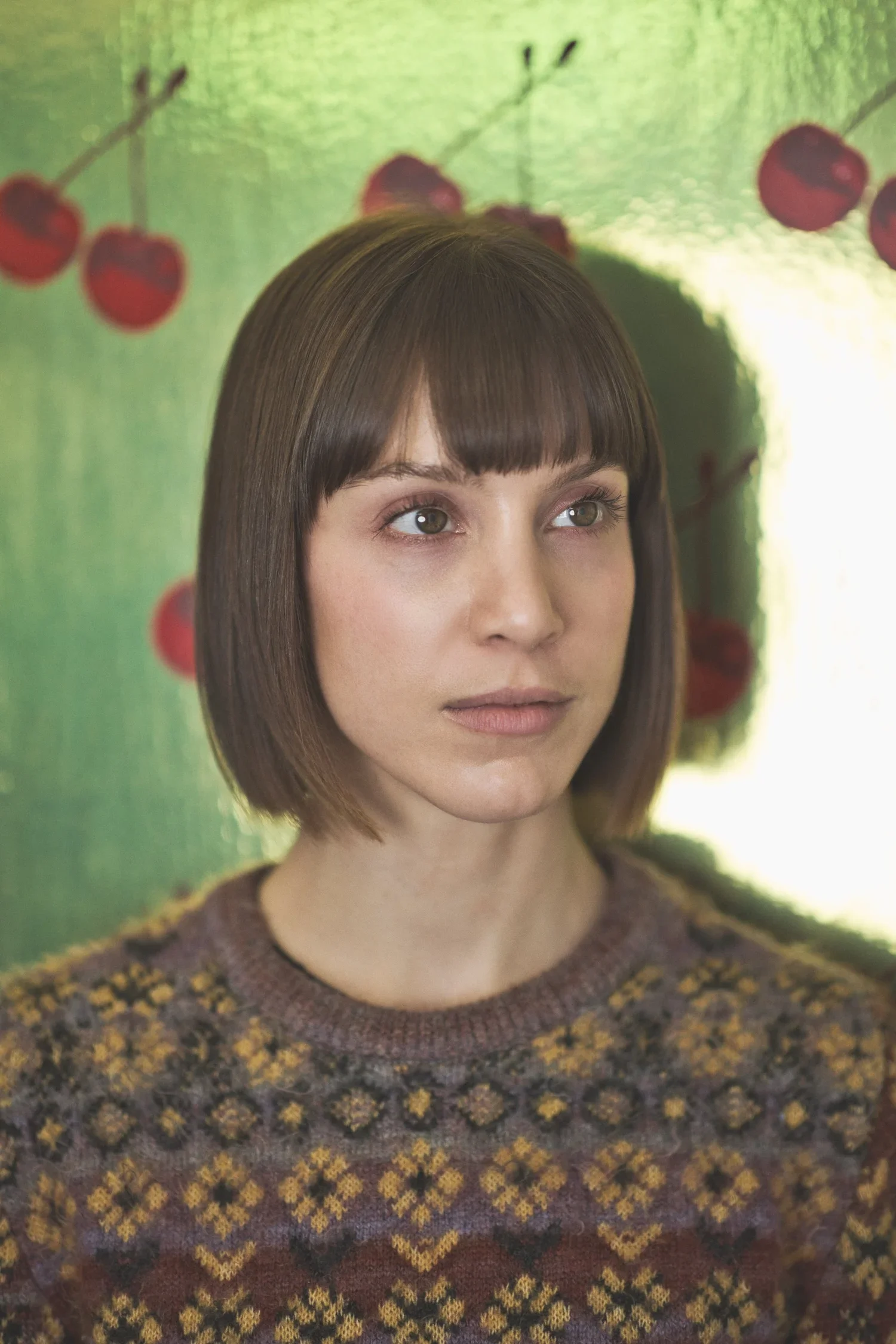 A woman with shoulder-length brown hair and gray eyes looking at the camera. She is wearing a patterned sweater with yellow, brown, and gray designs. The background features a green backdrop with red berries and green leaves.