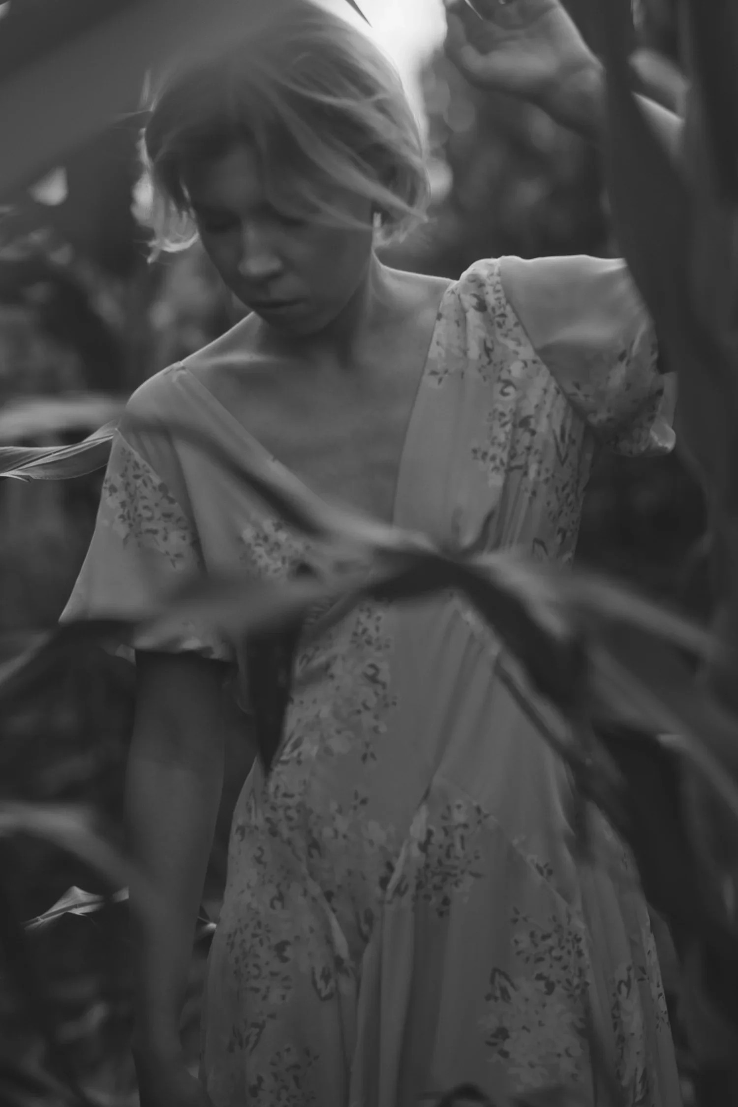 A woman with short blonde hair wearing a patterned dress is outdoors, surrounded by blurred foliage, with sunlight creating a halo effect around her head.
