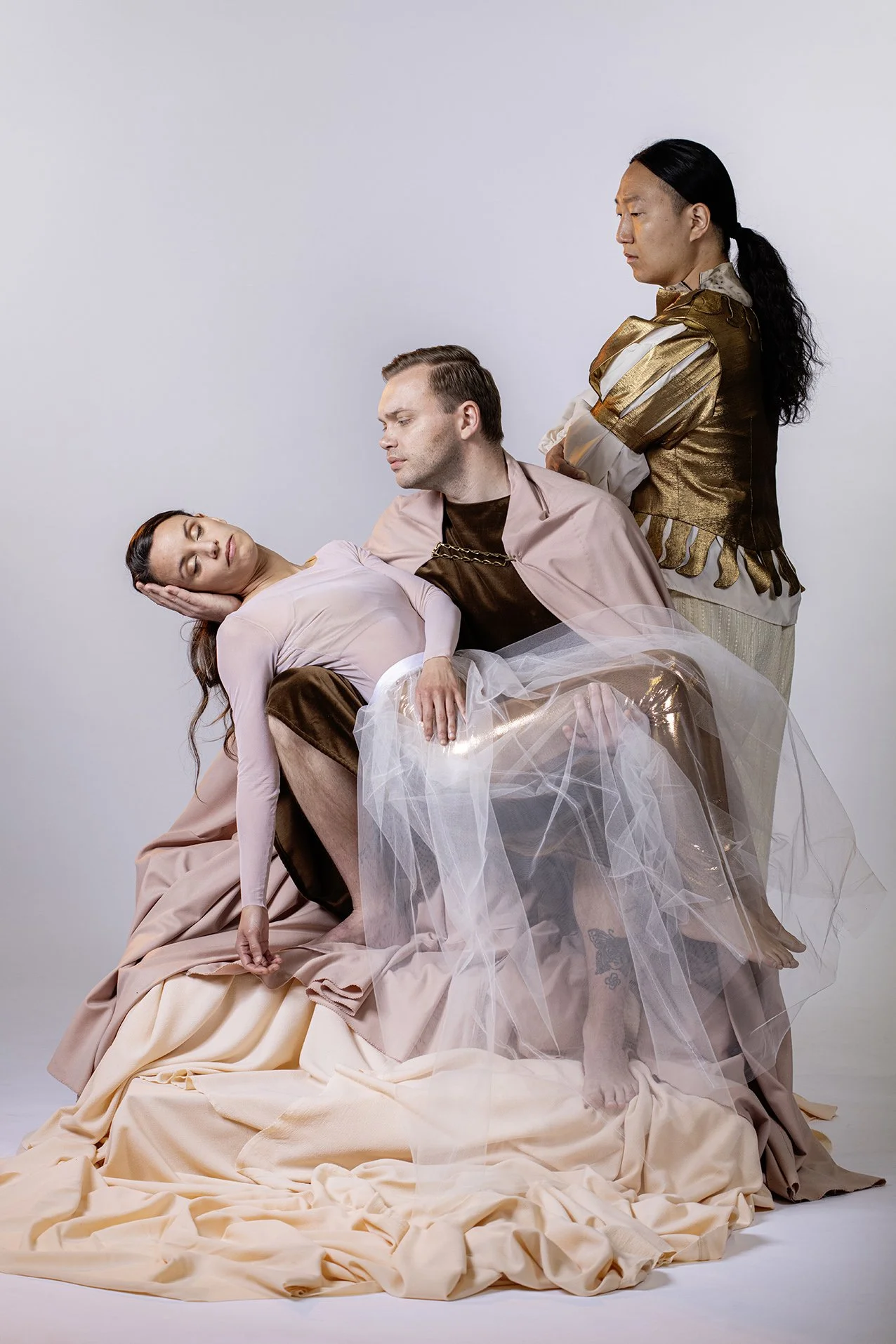 Three people, two men and one woman, are posed in a studio with a plain background. The woman, with dark hair, is reclining with her eyes closed, resting her head on the knee of the man in the middle, who has short hair and is looking at her. The woman is wearing a light pink dress, and the man is wearing a light pink jacket over a dark shirt. The third person, standing behind the seated pair, has long black hair tied back and is wearing a shiny gold and white outfit, looking downward with arms crossed.