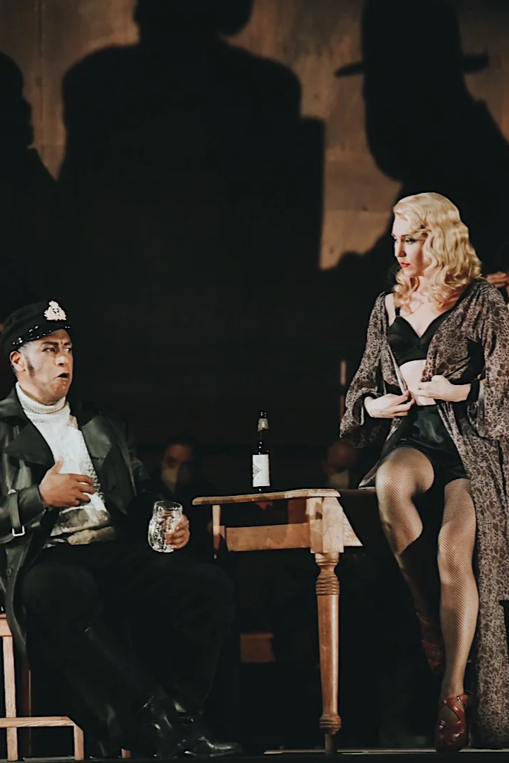 A theatrical scene with two actors: a man in a police uniform holding a glass and looking surprised, and a woman in provocative clothing sitting on a table with her legs crossed, holding a piece of paper with a bottle and a glass nearby, in a dimly lit setting.
