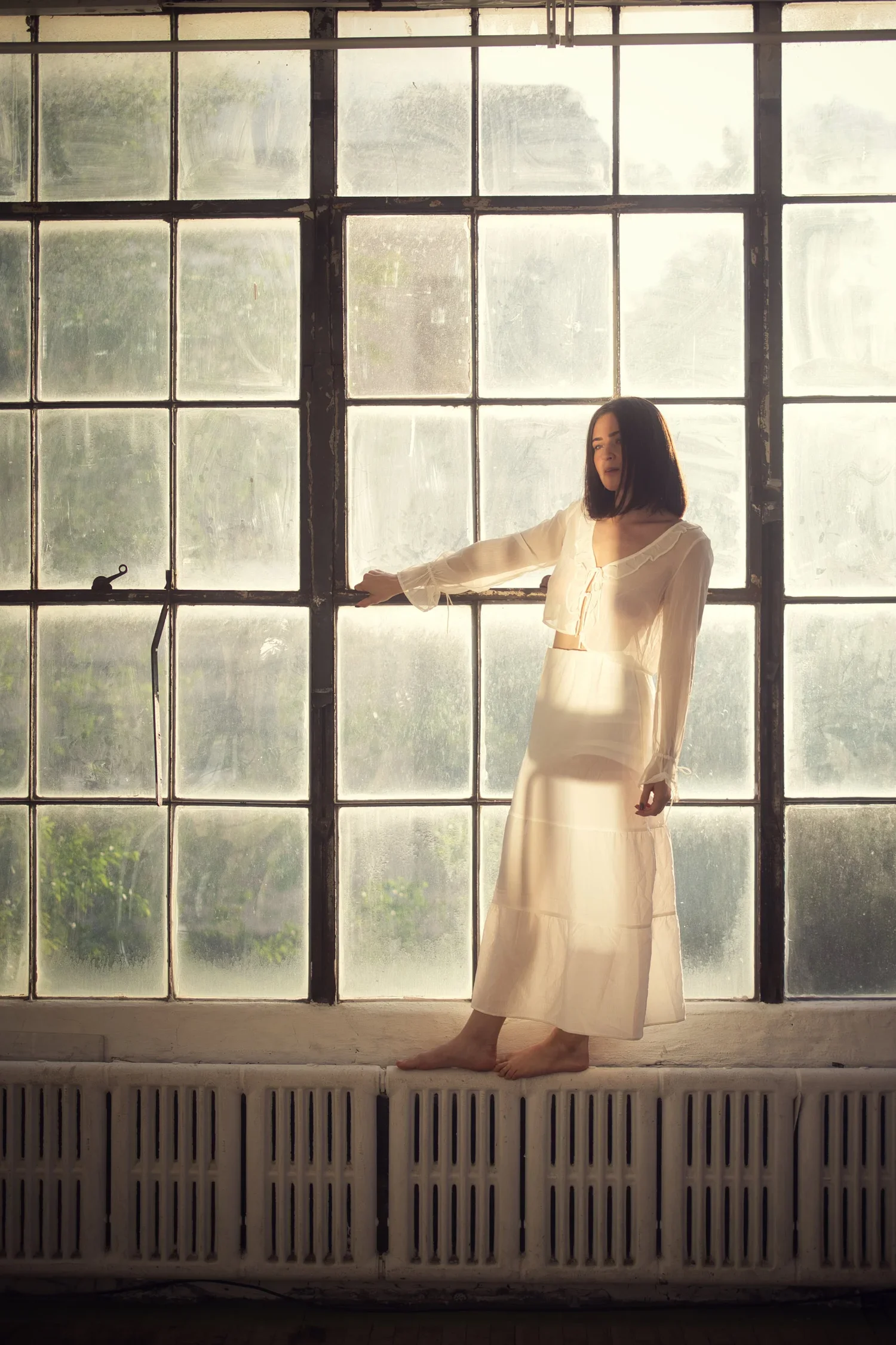 A woman in a white dress standing barefoot on a windowsill in front of large industrial-style windows with a view of greenery outside.
