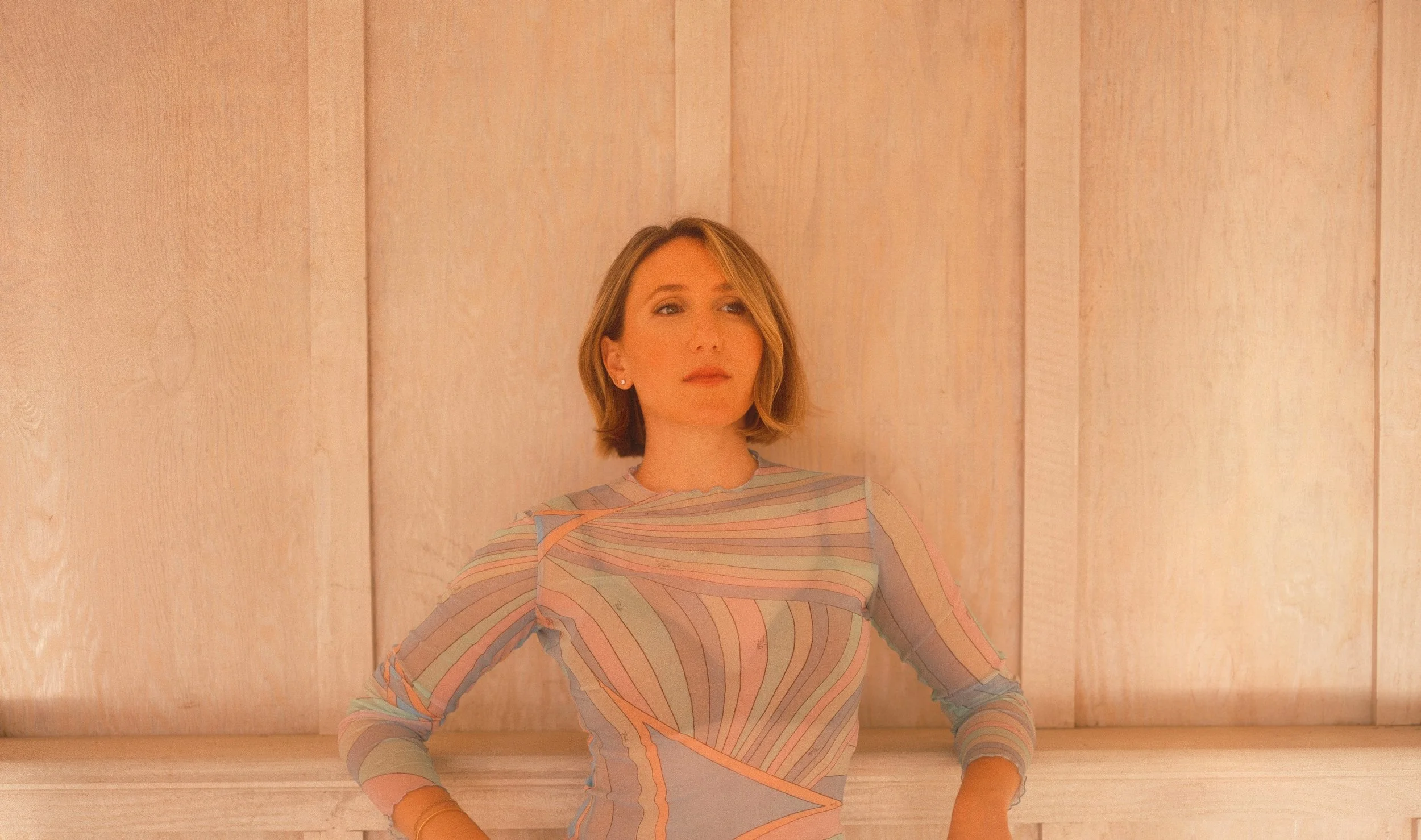 A woman with shoulder-length blonde hair, wearing a long-sleeved dress with pastel-colored wavy lines, standing against a wooden-paneled wall with her hands on her hips.