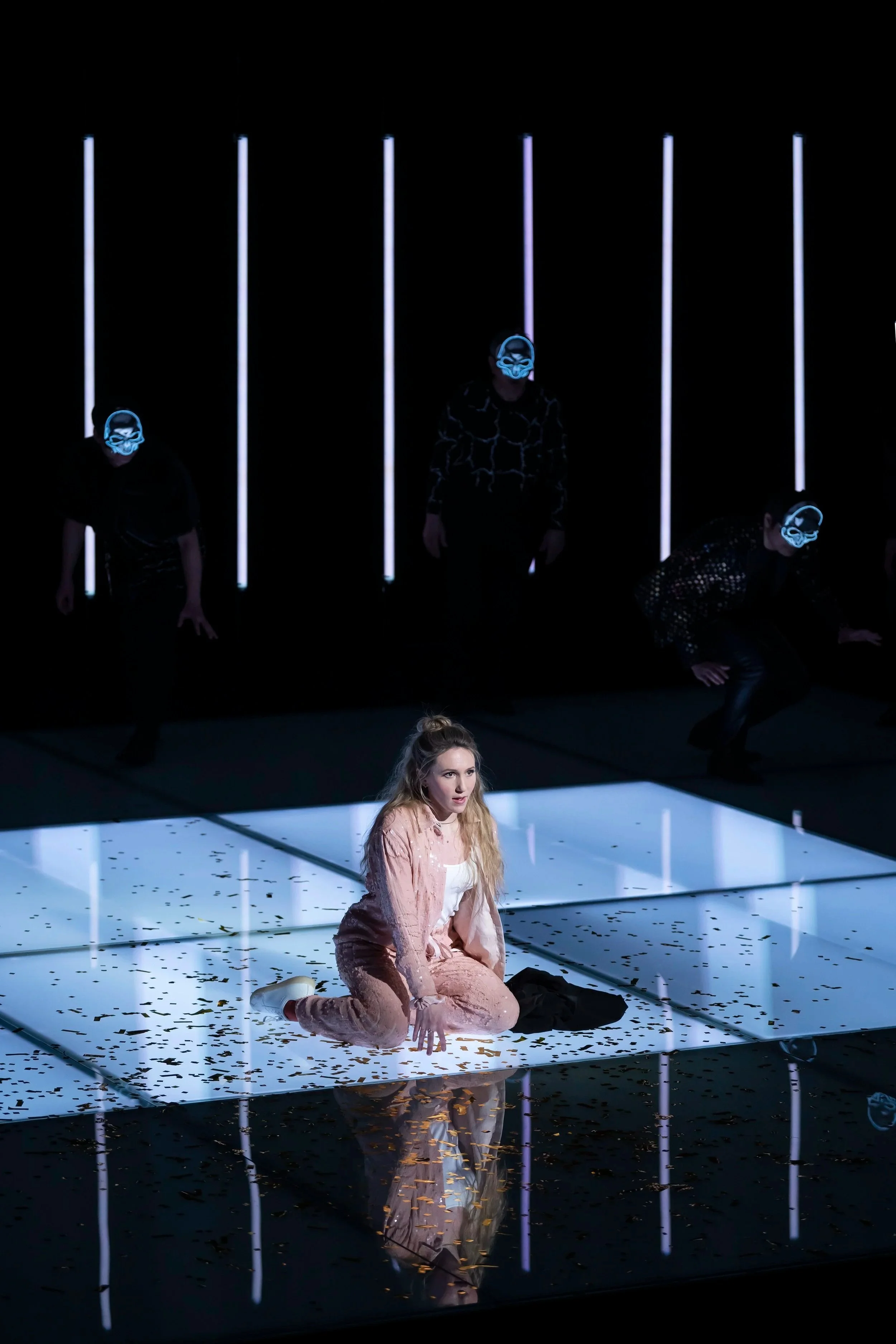 Performer kneeling on a reflective stage with scattered confetti and a mirror-like surface, wearing a pink velvet outfit, with behind a dark background featuring vertical neon lights and masked dancers wearing neon masks.