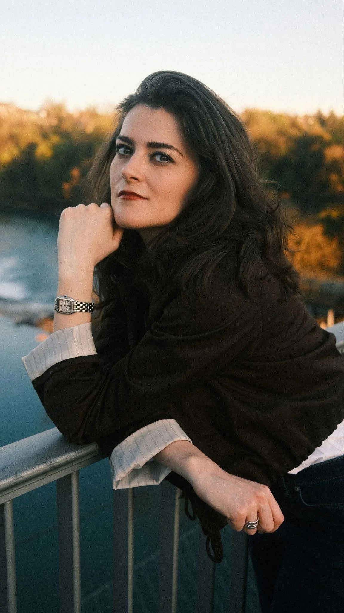 A woman with long dark hair and light skin leaning on a railing outdoors during sunset, wearing a black jacket, a watch, and a ring, with autumn trees and a river in the background.