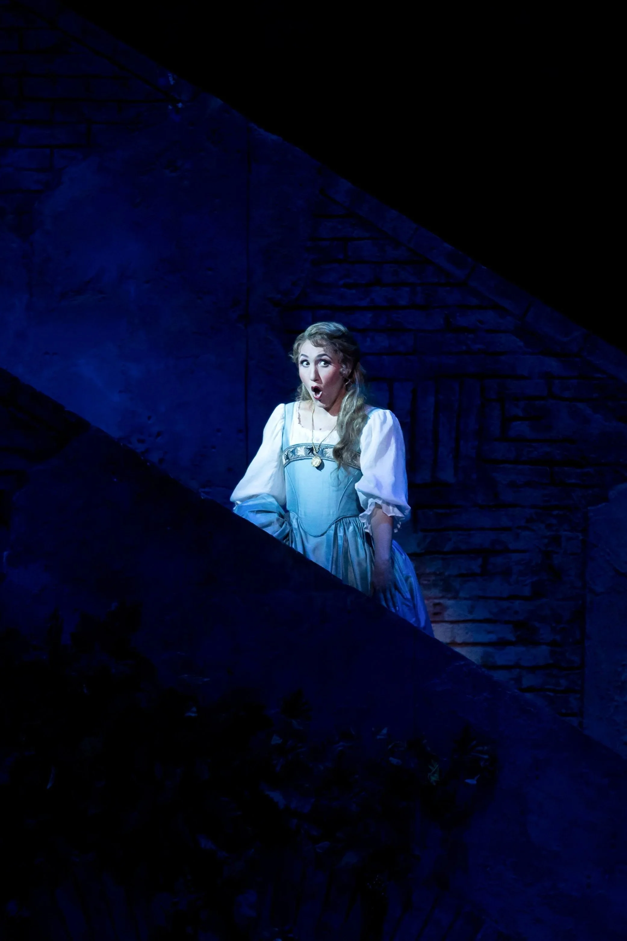 A woman dressed in historical costume with a surprised expression, performing on a dark stage with blue lighting and brick wall backdrop.