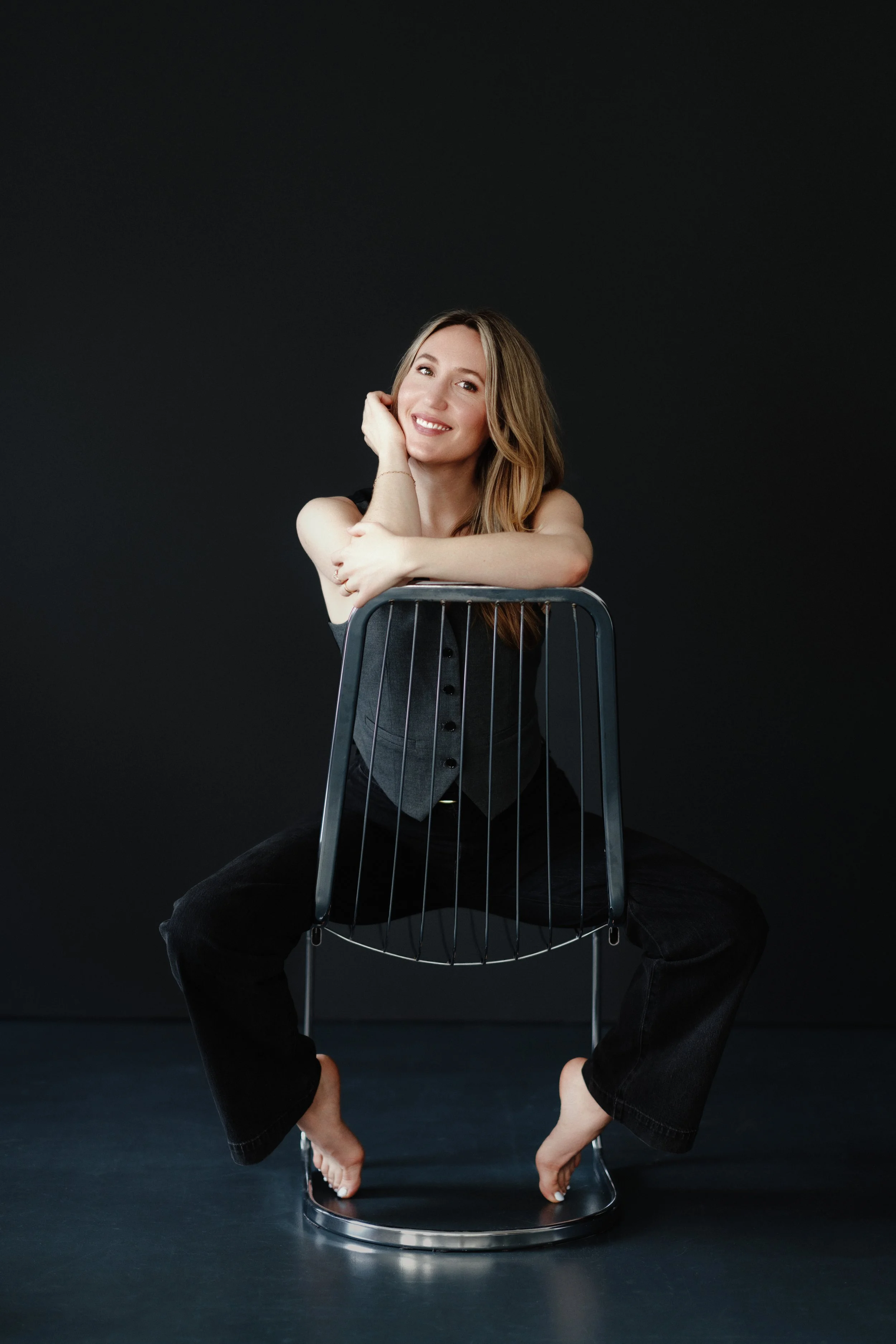 A smiling woman with long, wavy light brown hair, sitting behind a metal chair, with her legs stretched around the chair and her toes touching the ground, supporting her body weight. She rests her chin on her right hand, looking directly at the camera, against a solid black background.