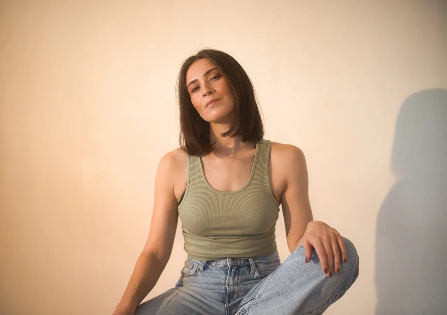 Young woman with shoulder-length brown hair sitting against a plain light-colored wall, wearing a green tank top and light wash jeans, looking at the camera with a relaxed expression.