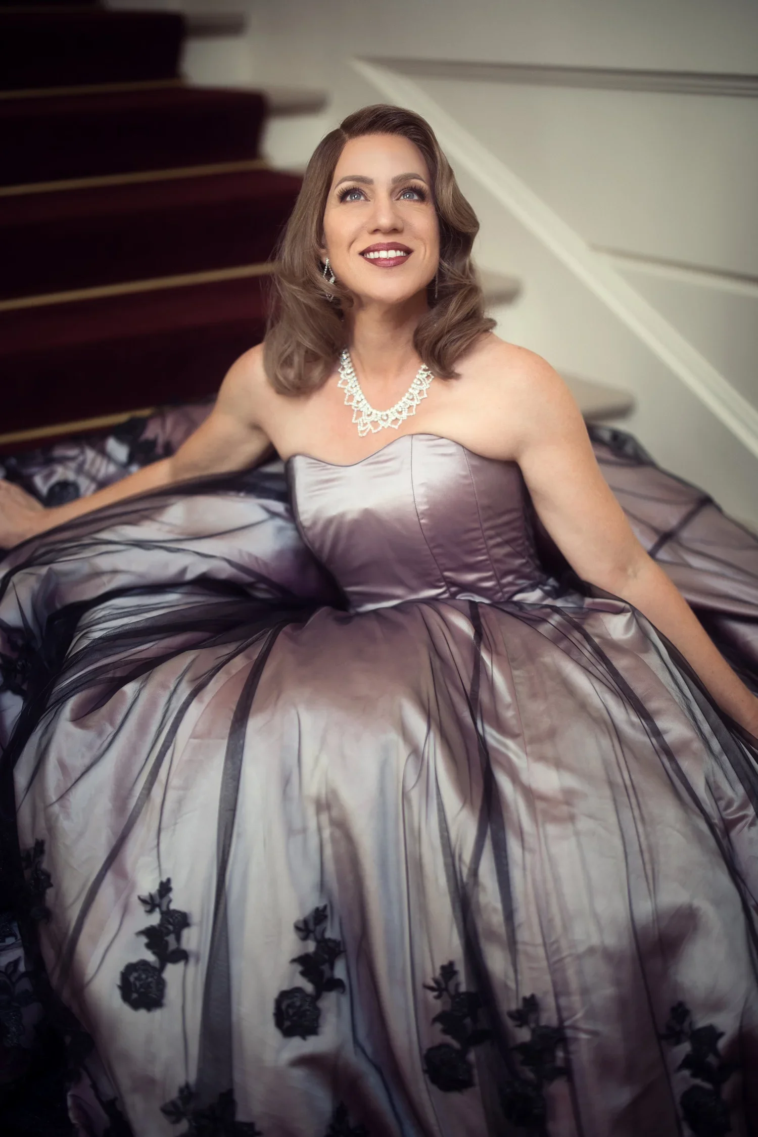 A woman with brown hair and blue eyes, wearing a strapless silver gown with black floral embroidery, a diamond necklace, and earrings, sitting on a bed with her head tilted slightly upward and smiling.