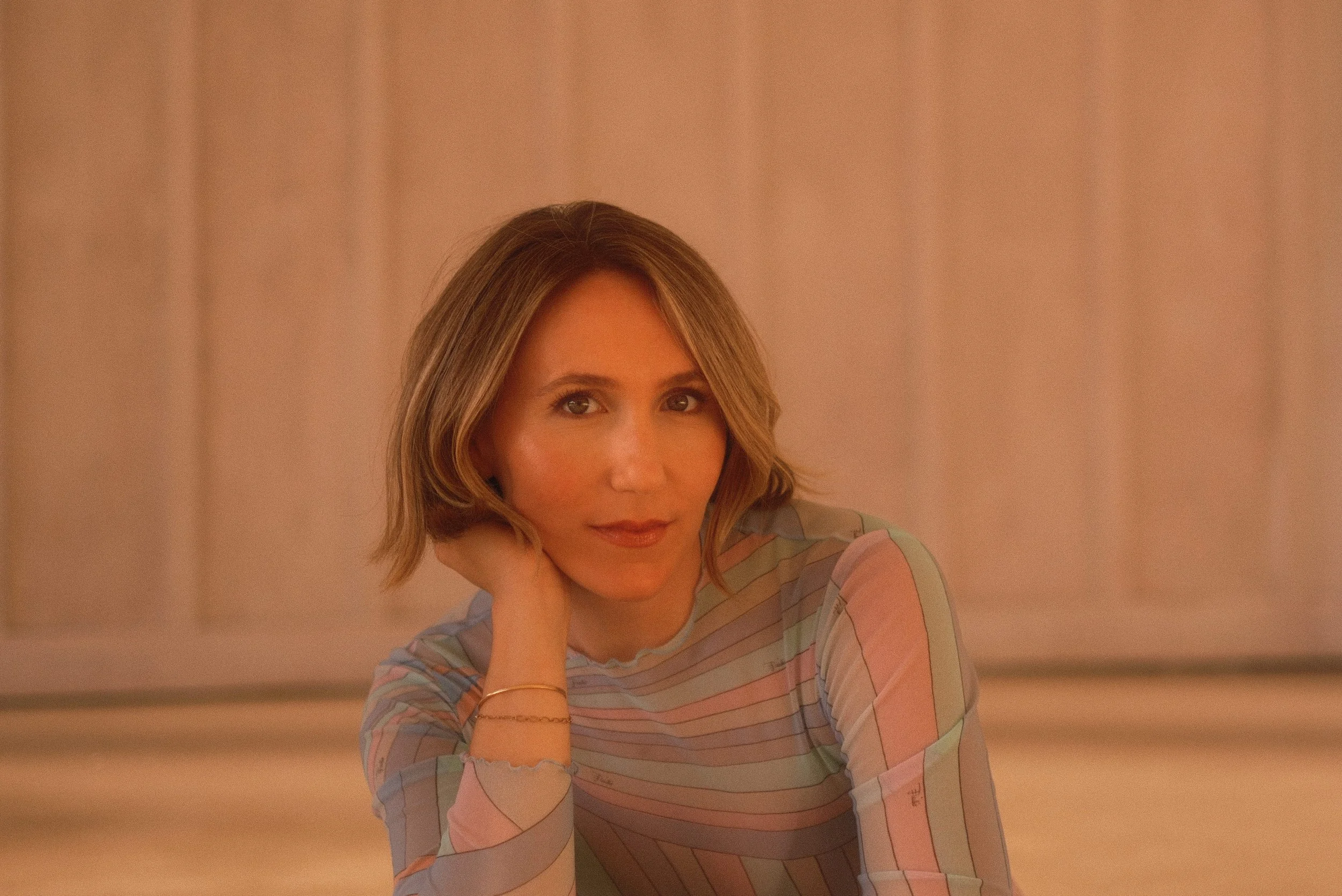 A woman with light brown hair styled in a shoulder-length bob, wearing a long-sleeved pastel-colored striped shirt, sitting against a soft coral-colored background, resting her chin on her hand, and looking directly at the camera.