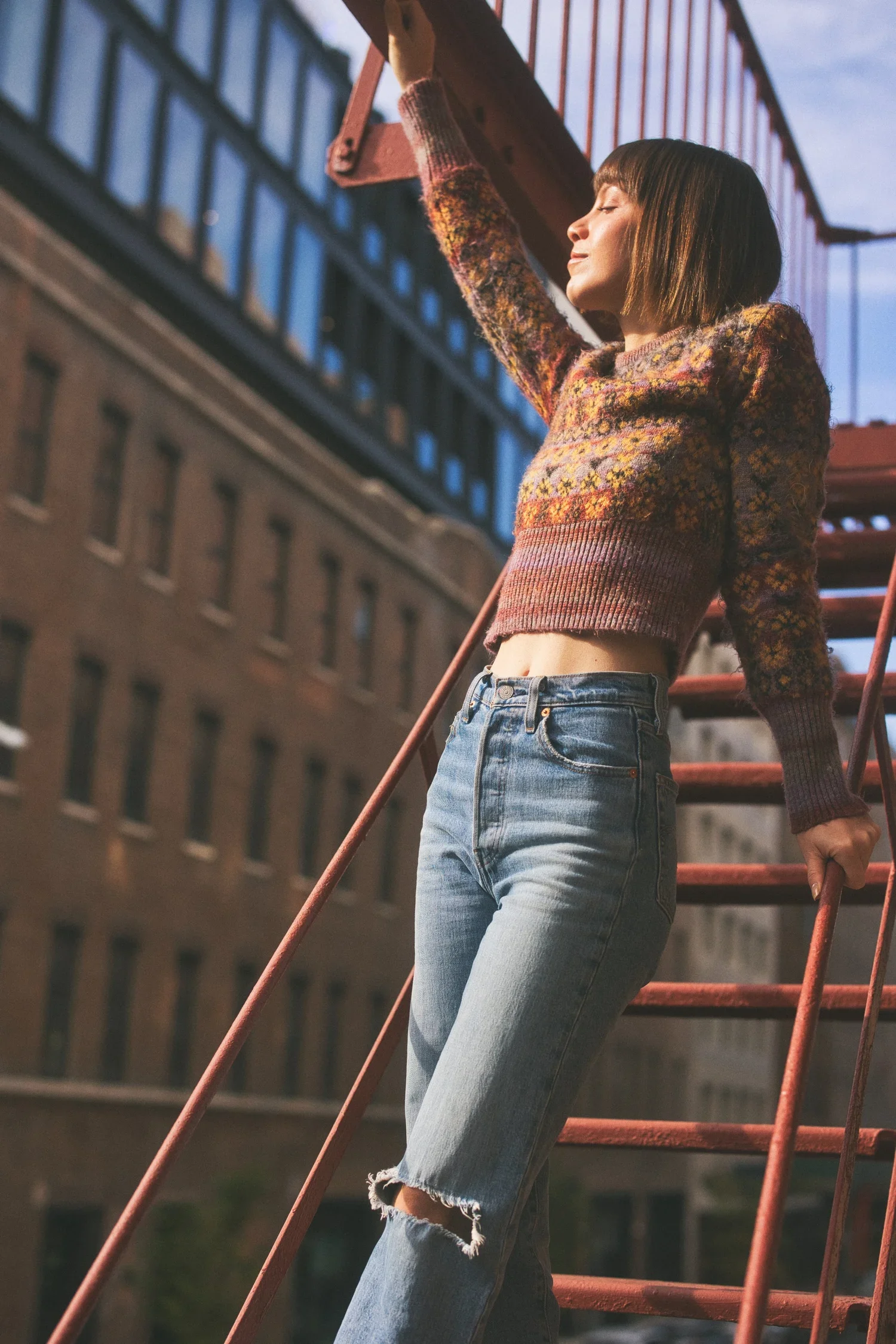 Young woman with a bob haircut and closed eyes, wearing a colorful knit sweater and ripped jeans, standing on an outdoor fire escape during sunset.