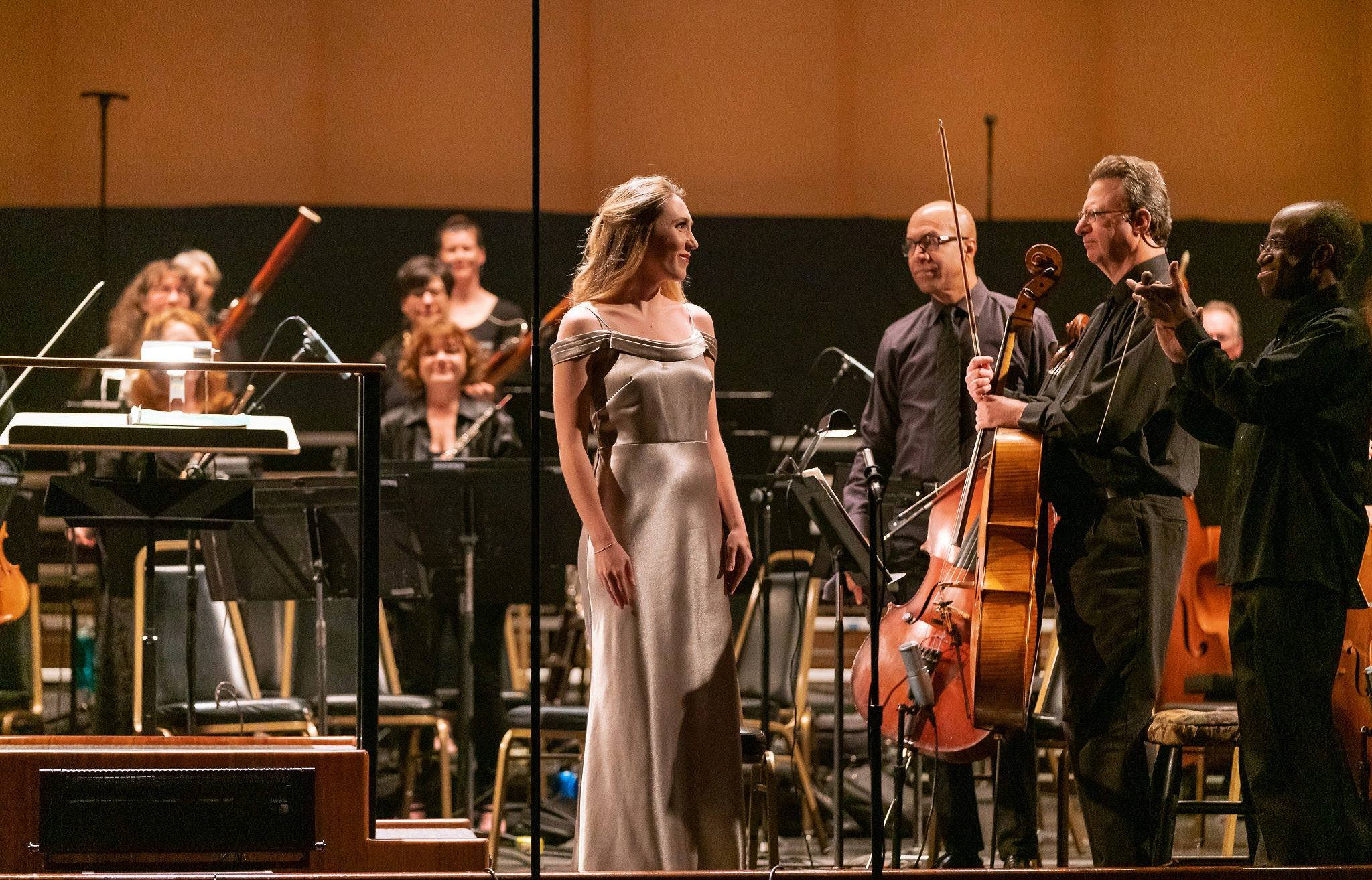 A woman in a satin dress stands on stage in front of musicians, holding a violin, during a concert.