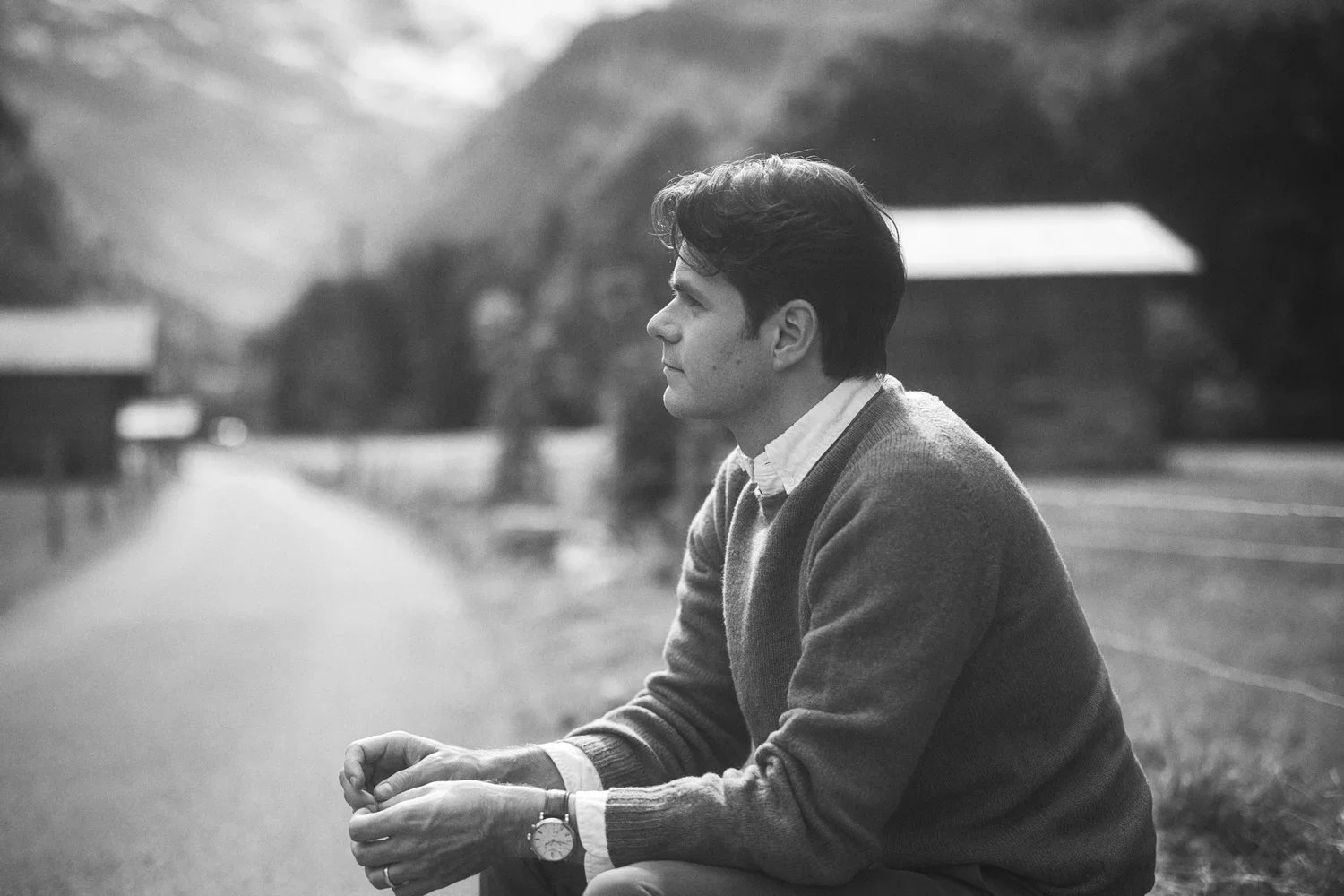 A young man with dark hair, wearing a sweater and a watch, sitting outdoors on a bench or step, looking contemplative, in a black-and-white photograph.