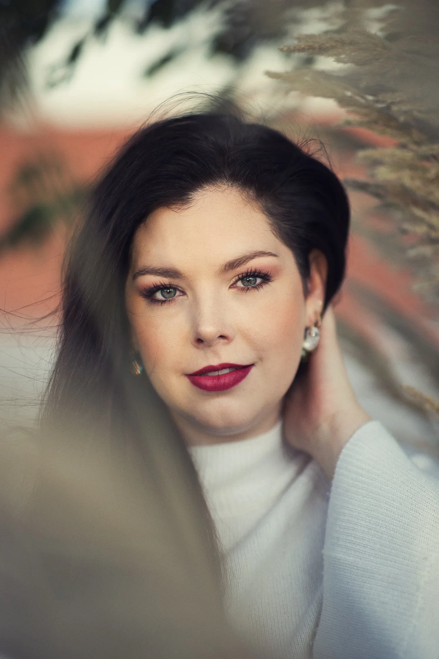 A woman with dark hair and blue eyes, wearing red lipstick and earrings, dressed in a white sweater, with a blurred background.